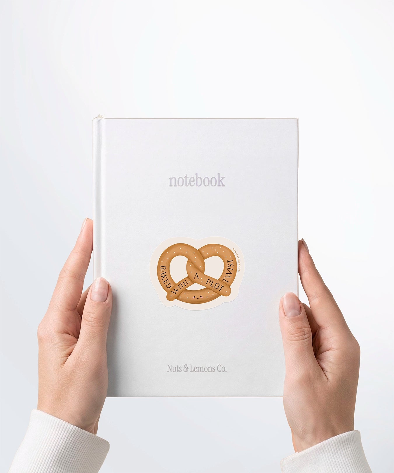 A pair of hands holding a white notebook with a Nuts & Lemons Co. diecut sticker on it featuring a pretzel with the phrase “Baked with a Plot Twist”