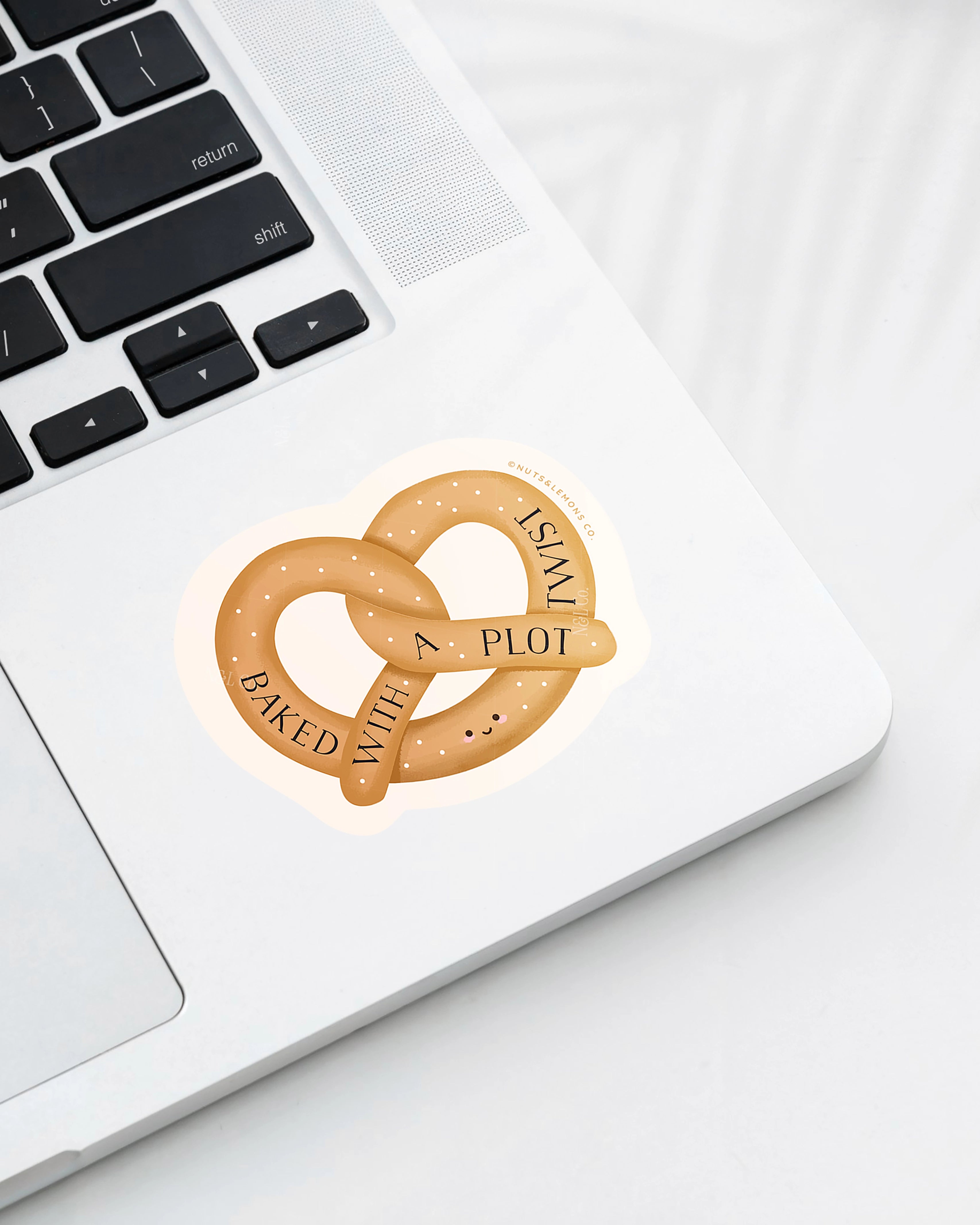 Laptop with a Nuts & Lemons Co. diecut sticker featuring a pretzel with the phrase “Baked with a Plot Twist”