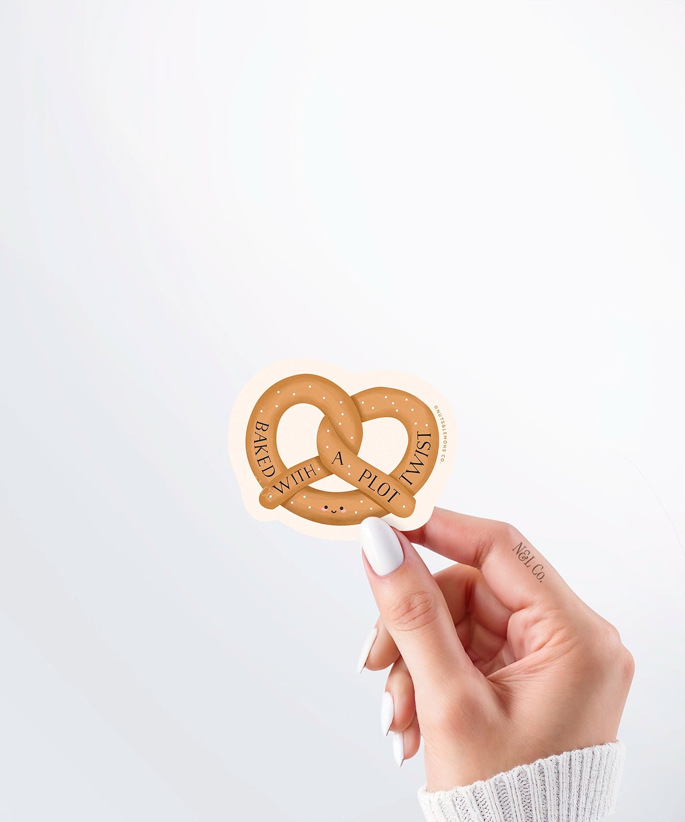 Hand holding a Nuts & Lemons Co. diecut sticker featuring a pretzel with the phrase “Baked with a Plot Twist”