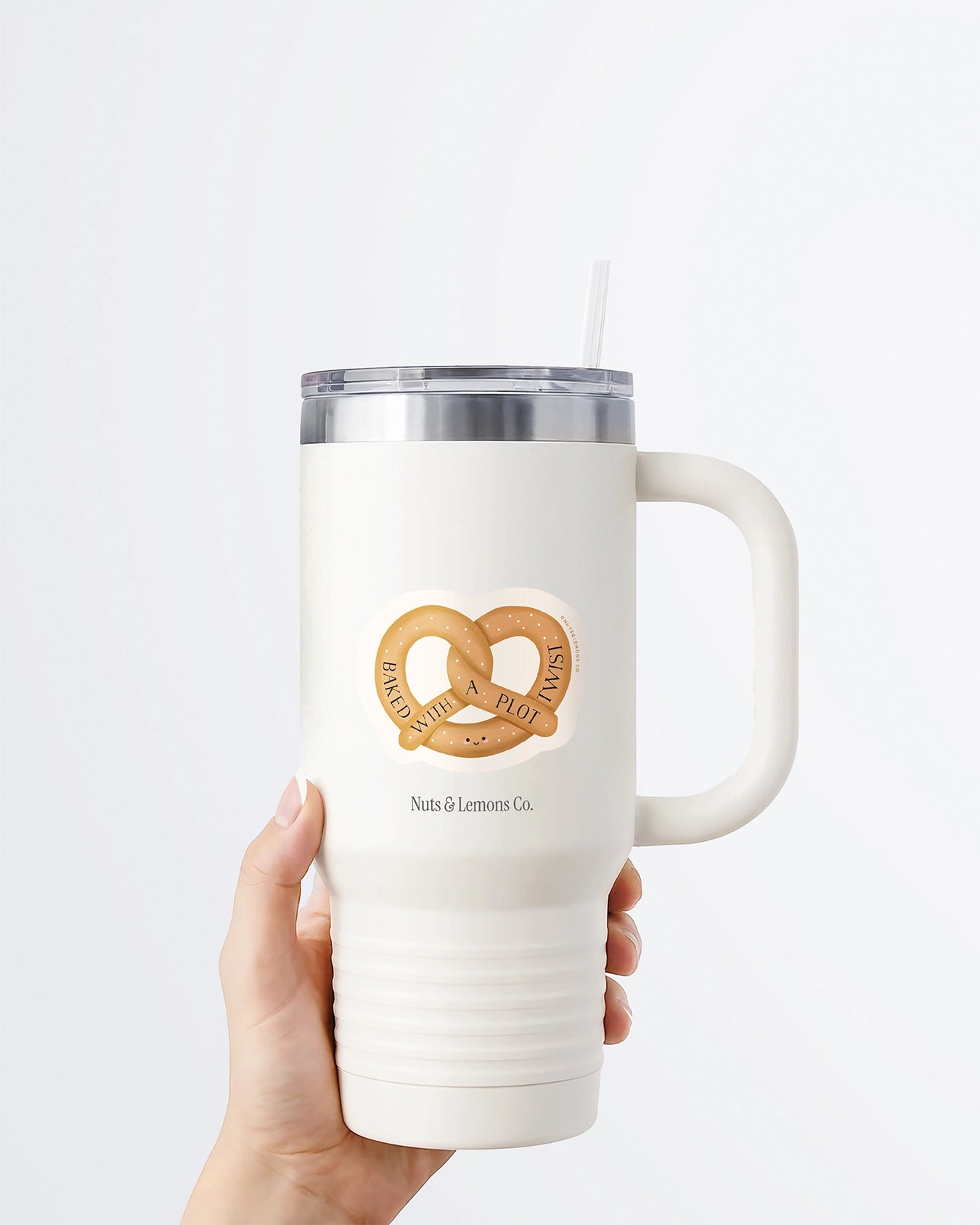 A hand holding a white tumbler with a Nuts & Lemons Co. diecut sticker on it featuring a pretzel with the phrase “Baked with a Plot Twist”