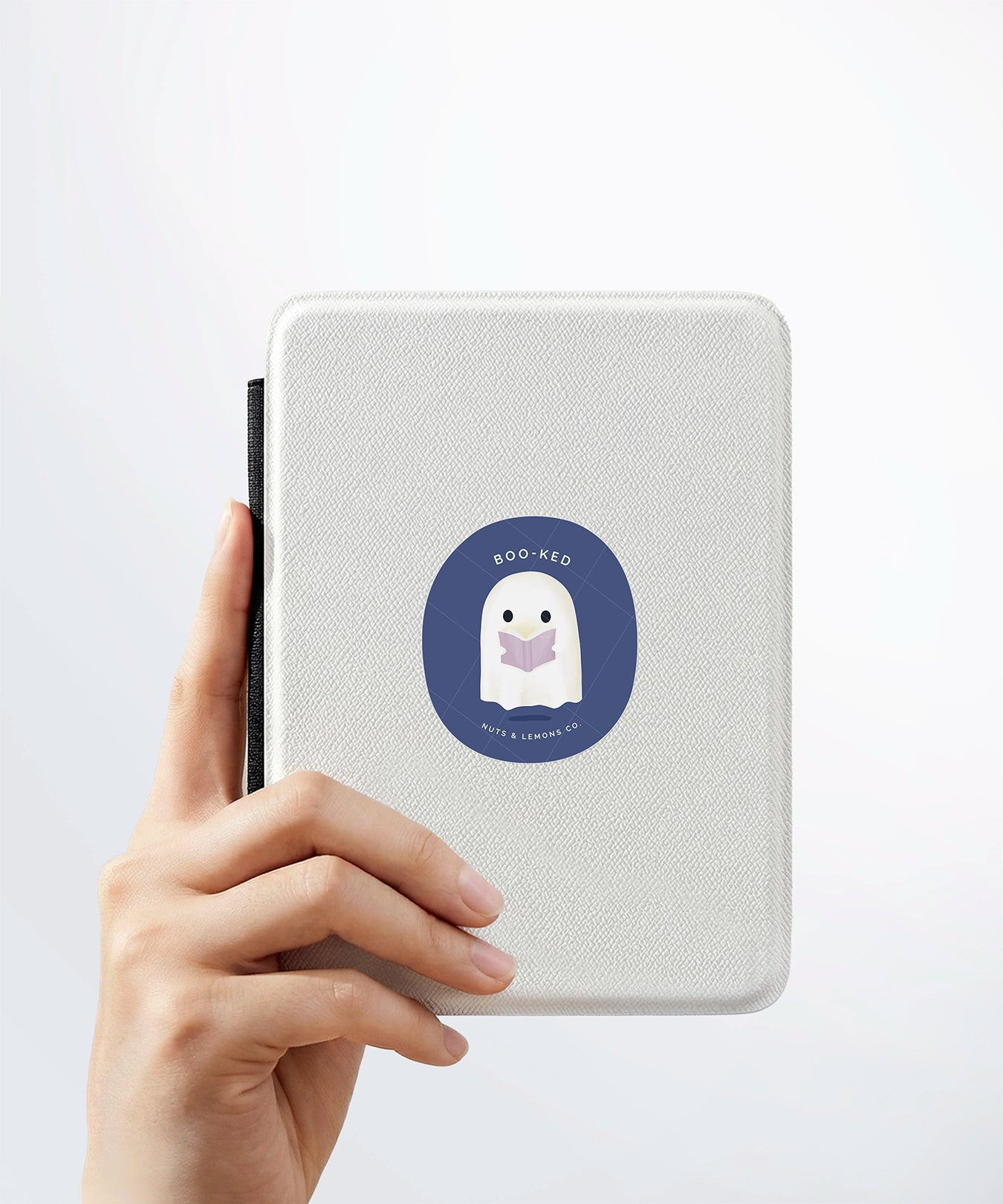 A hand holding a white kindle cover with a Nuts & Lemons Co. diecut sticker on it featuring a cute ghostie  reading a glowing book with the phrase “Boo-ked”