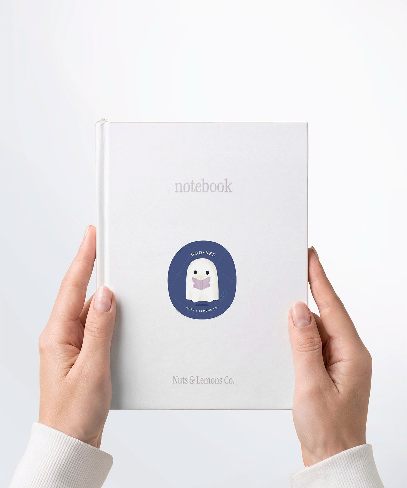A pair of hands holding a white notebook with a Nuts & Lemons Co. diecut sticker on it featuring a cute ghostie  reading a glowing book with the phrase “Boo-ked”