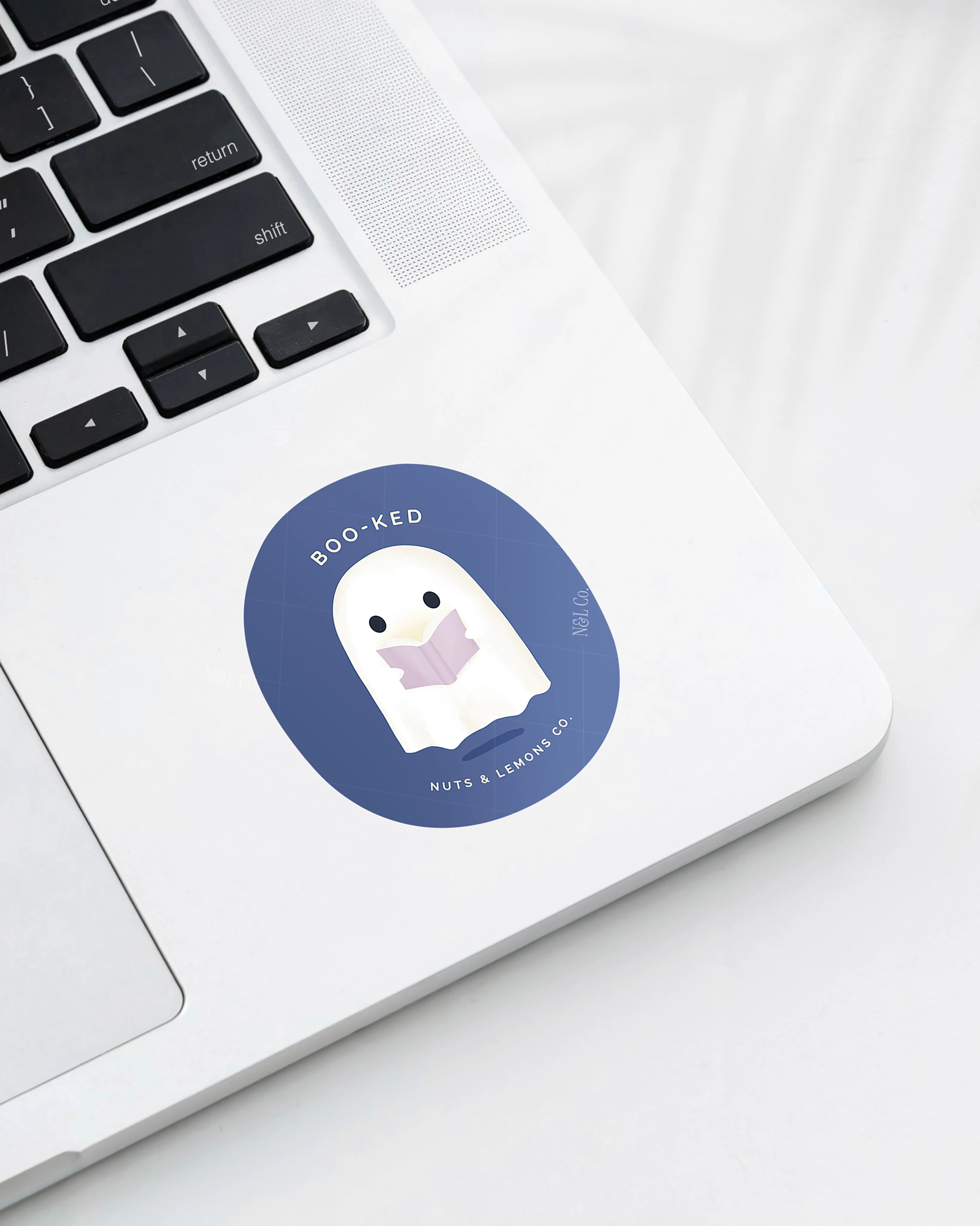 Laptop displaying a Nuts & Lemons Co. diecut sticker featuring a cute ghostie  reading a glowing book with the phrase “boo-ked”
