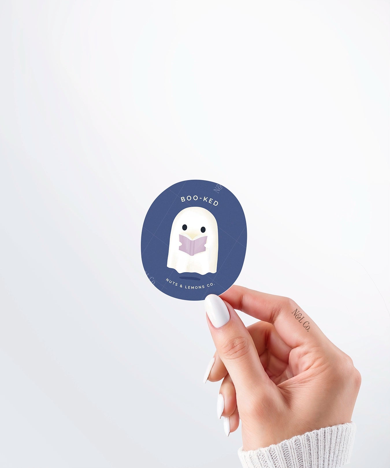 Hand holding a Nuts & Lemons Co. diecut sticker featuring a cute ghostie  reading a glowing book with the phrase “boo -ked”