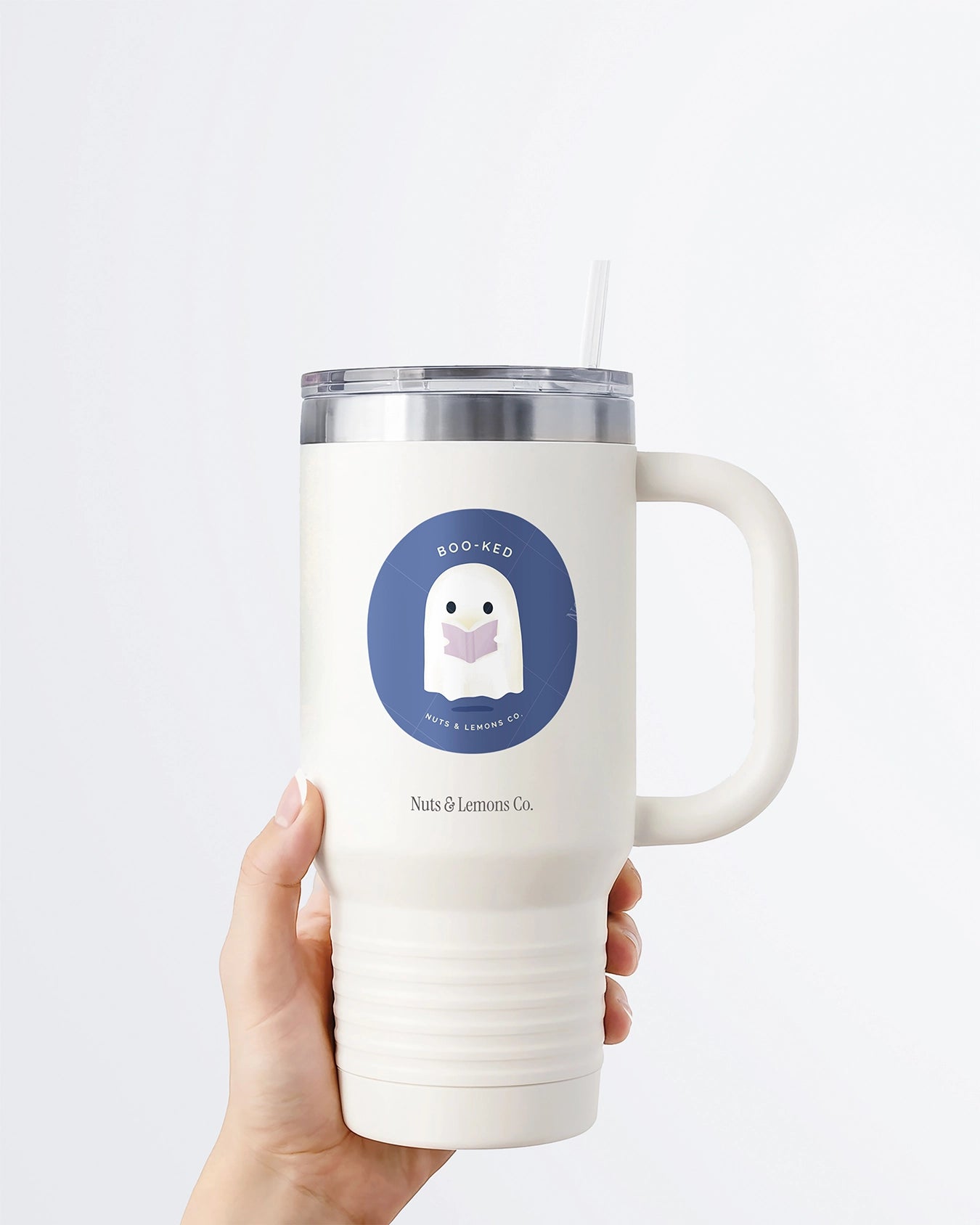 Hand holding a tumbler displaying Nuts & Lemons Co. diecut sticker featuring  a cute ghostie  reading a glowing book with the phrase “boo-ked”