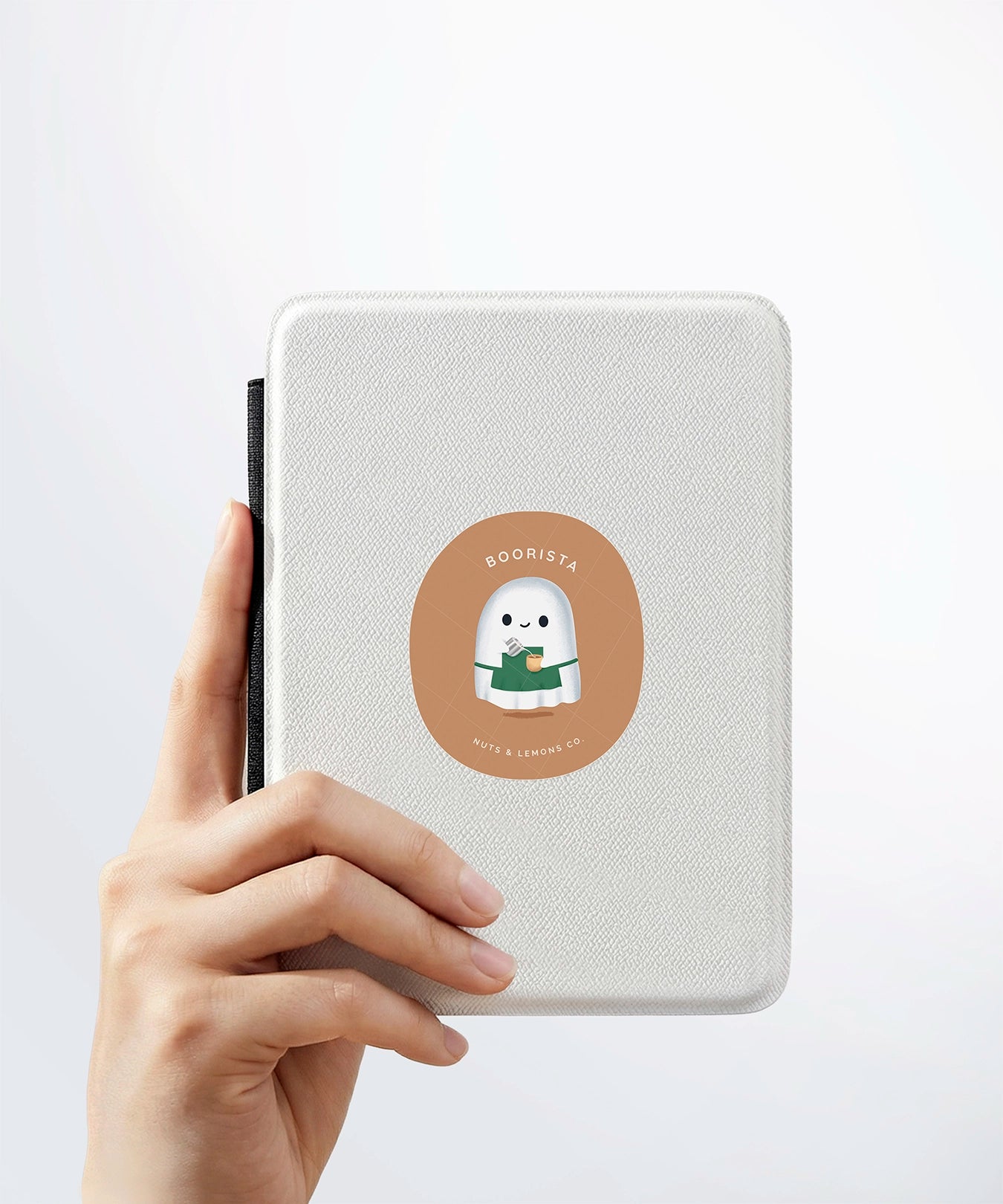 A hand holding a white kindle cover with a Nuts & Lemons Co. diecut sticker on it featuring a cute ghostie barista making a latte with the phrase “Boo-rista”
