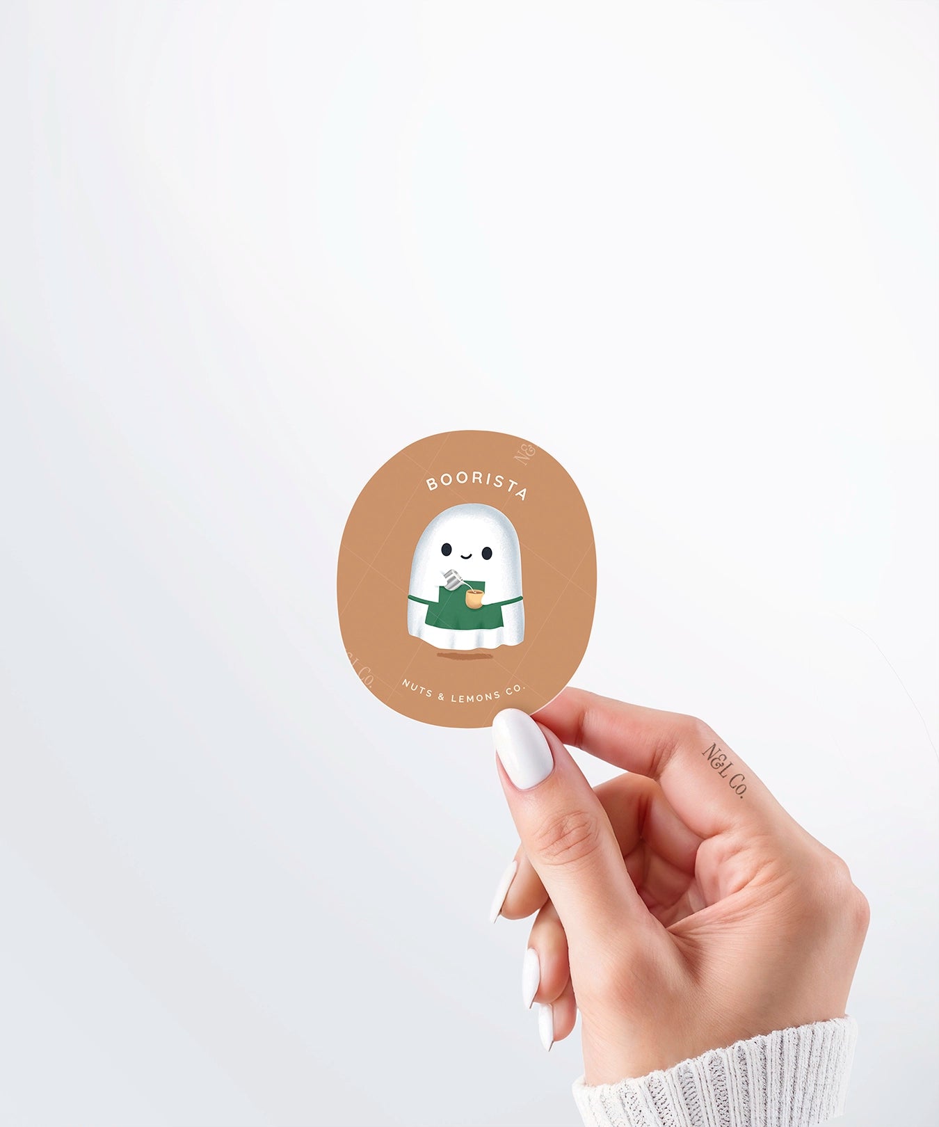 Hand holding a Nuts & Lemons Co. diecut sticker featuring a cute ghostie barista making a latte  with the phrase “boorista”