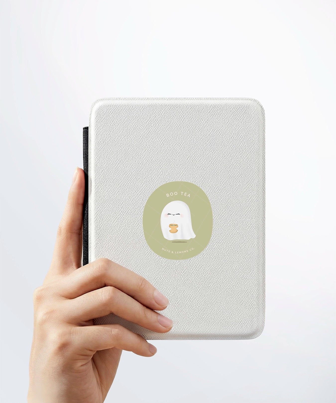 A hand holding a white kindle cover with a Nuts & Lemons Co. diecut sticker on it featuring a cute ghostie holding hot green tea with the phrase “Boo-tea”