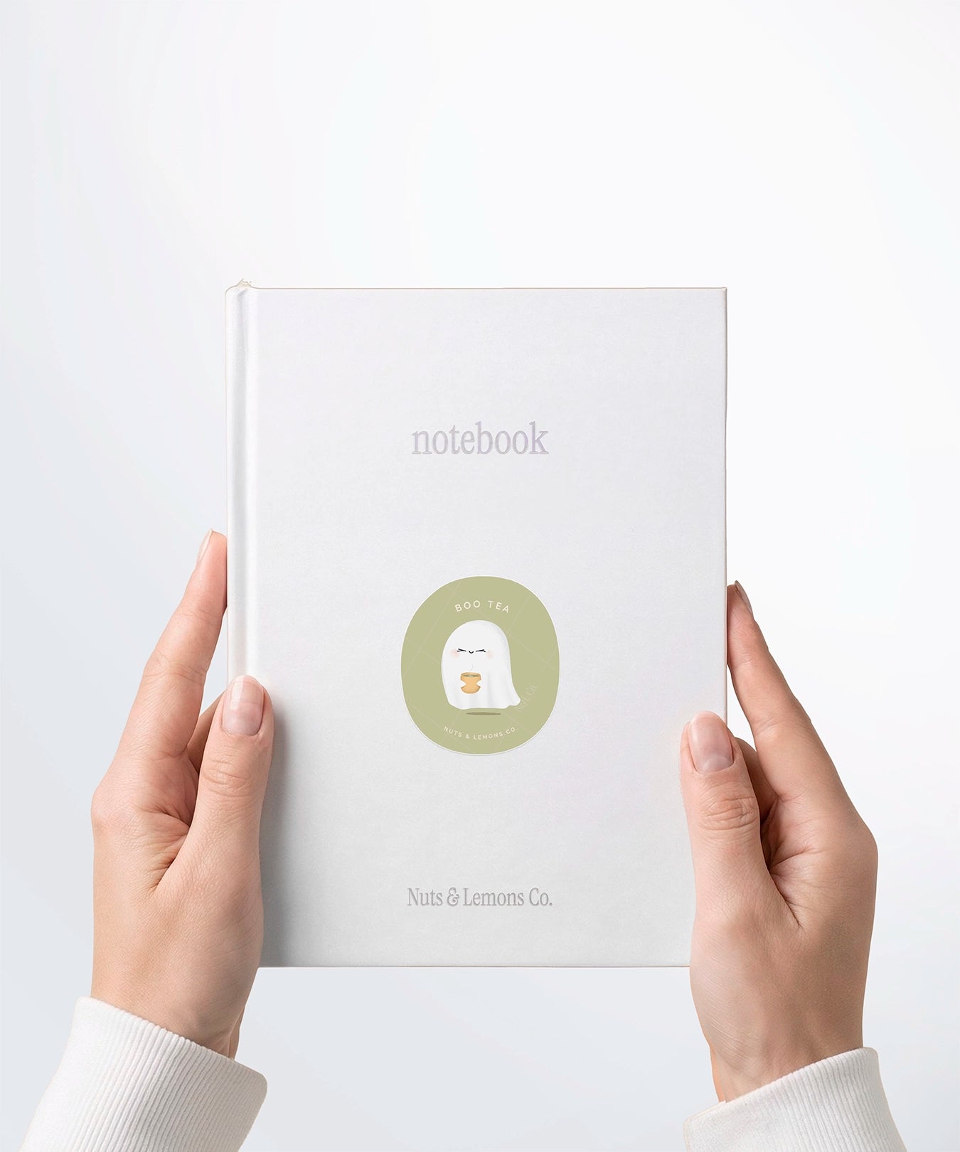 A pair of hands holding a white notebook with a Nuts & Lemons Co. diecut sticker on it featuring a cute ghostie holding hot green tea with the phrase “Boo-tea”