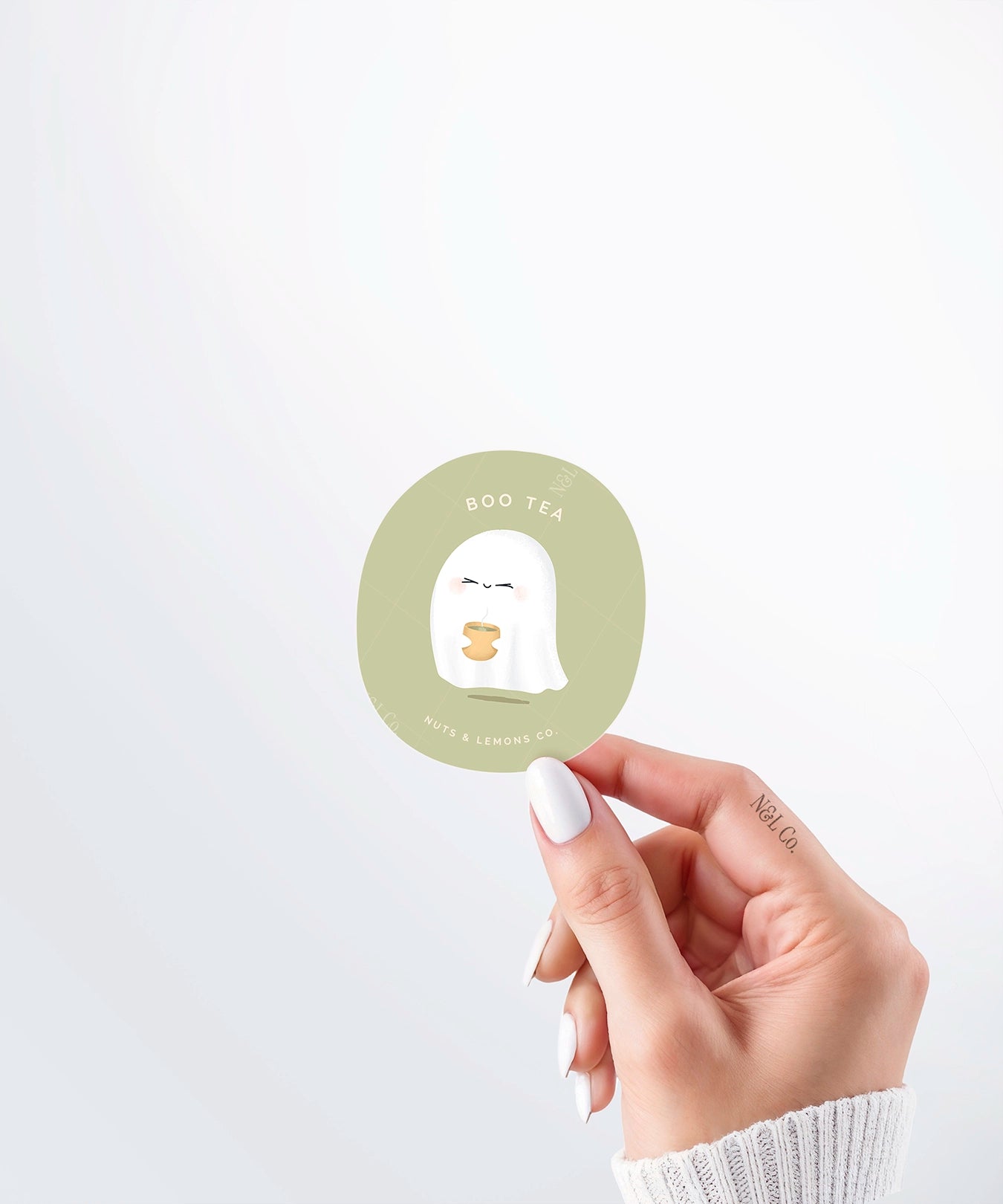 Hand holding a Nuts & Lemons Co. diecut sticker featuring a cute ghostie holding hot green tea with the phrase “boo tea”