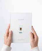 A pair of hands holding a white notebook with a Nuts & Lemons Co. diecut sticker on it featuring a polar bear chomping a brick of chocolate with the phrase “Miner”