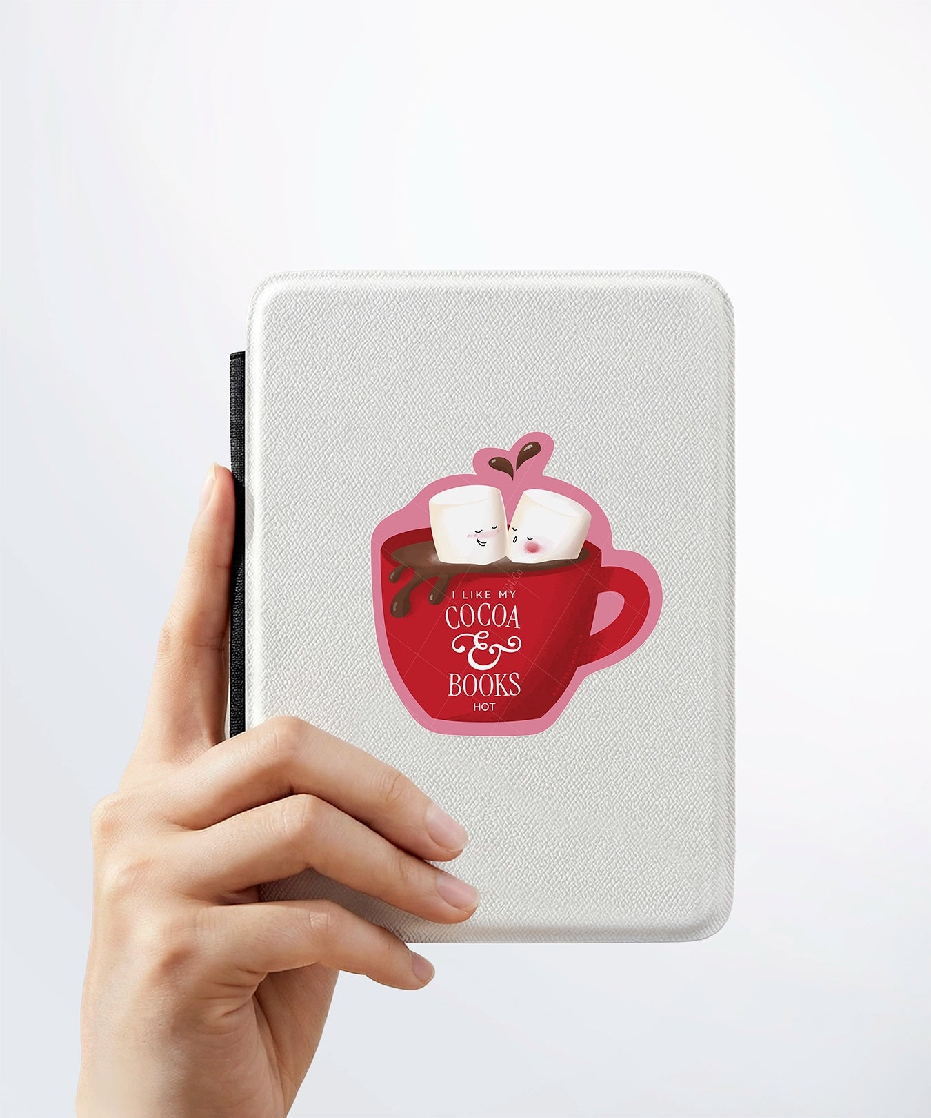 A hand holding a white kindle cover with a Nuts & Lemons Co. diecut sticker on it featuring a mug of hot cocoa with two lover marshmallows with the phrase “I Like My Cocoa & Books Hot"