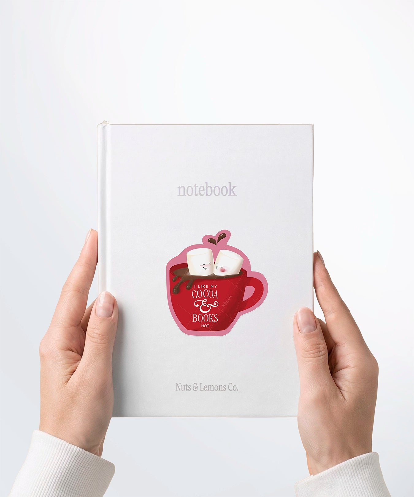 A pair of hands holding a white notebook with a Nuts & Lemons Co. diecut sticker on it featuring a mug of hot cocoa with two lover marshmallows with the phrase “I Like My Cocoa & Books Hot"