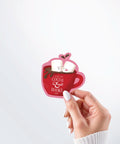 Hand holding a Nuts & Lemons Co. diecut sticker featuring a mug of hot cocoa with two marshmallows getting spicy with the phrase “I like my cocoa & books hot”
