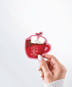 Hand holding a Nuts & Lemons Co. diecut sticker featuring a mug of hot cocoa with two marshmallows getting spicy with the phrase “I like my cocoa & books hot”