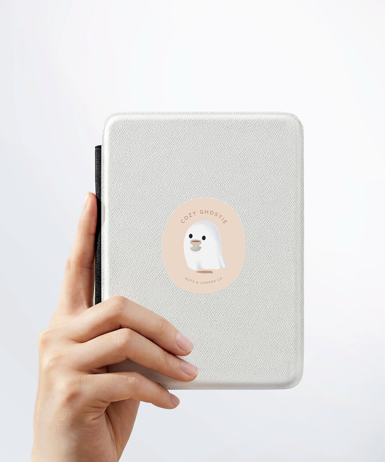A hand holding a white kindle cover with a Nuts & Lemons Co. diecut sticker on it featuring a cute ghostie holding a hot beverage with the phrase “Cozy Ghostie”
