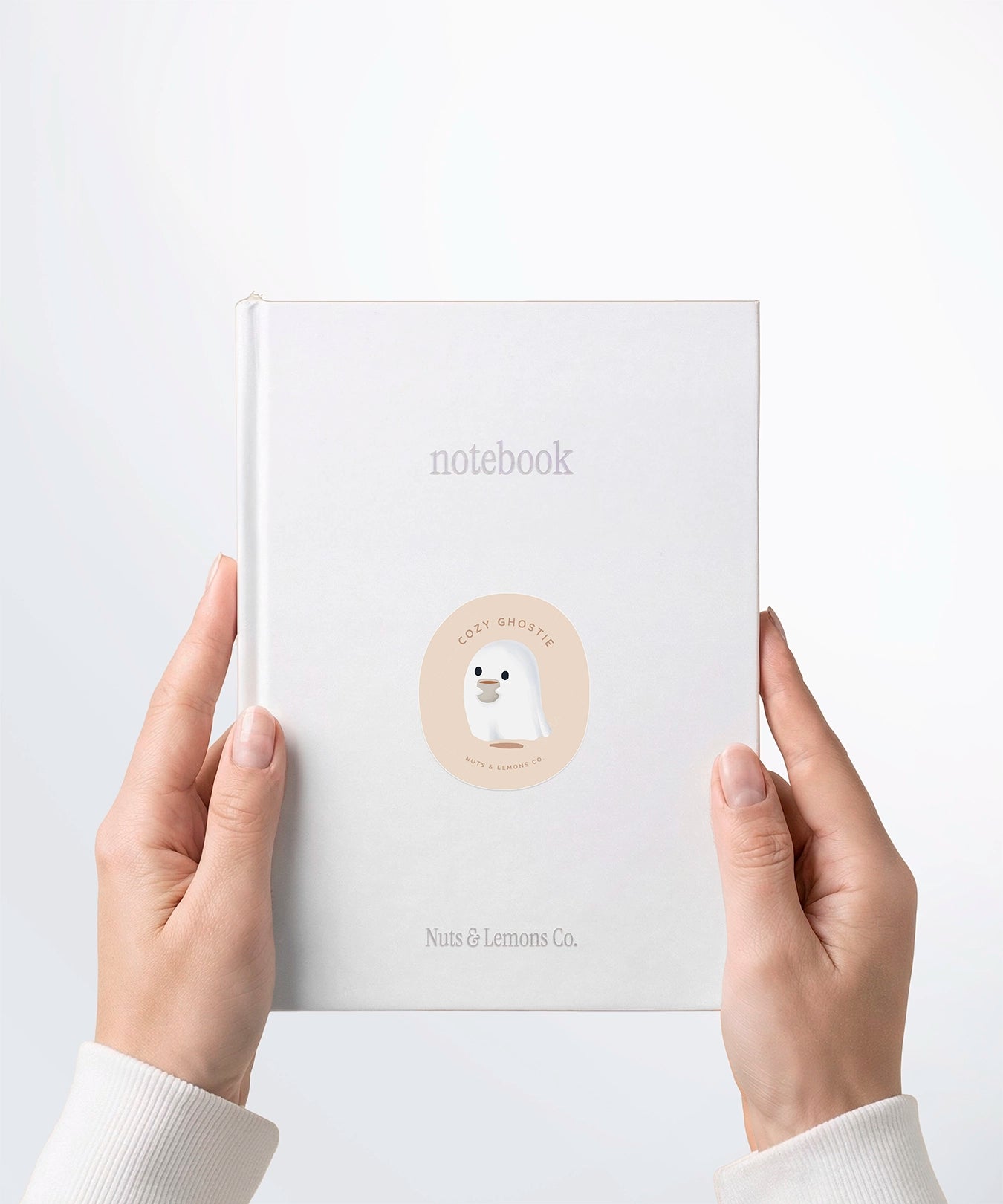 A pair of hands holding a white notebook with a Nuts & Lemons Co. diecut sticker on it featuring a cute ghostie holding a hot beverage with the phrase “Cozy Ghostie”