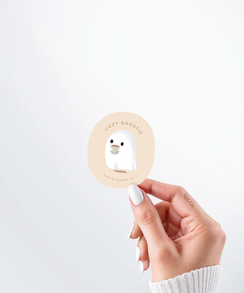 Hand holding a Nuts & Lemons Co. diecut sticker featuring a cute ghostie holding a hot beverage with the phrase “cozy ghostie”
