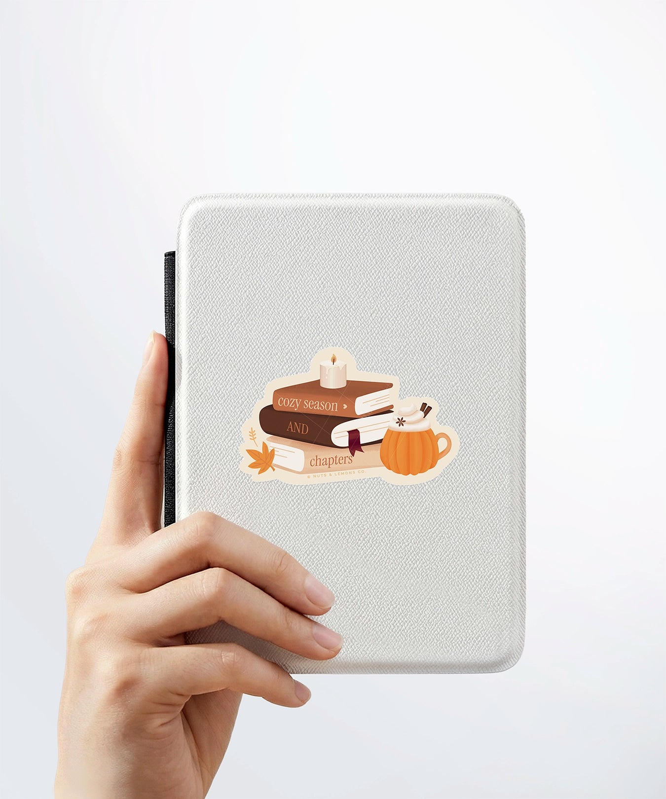 A hand holding a white kindle cover with a Nuts & Lemons Co. diecut sticker on it featuring a stack of autumnal books, candle and pumpkin spice latte with the phrase “Cozy Season and Chapters”