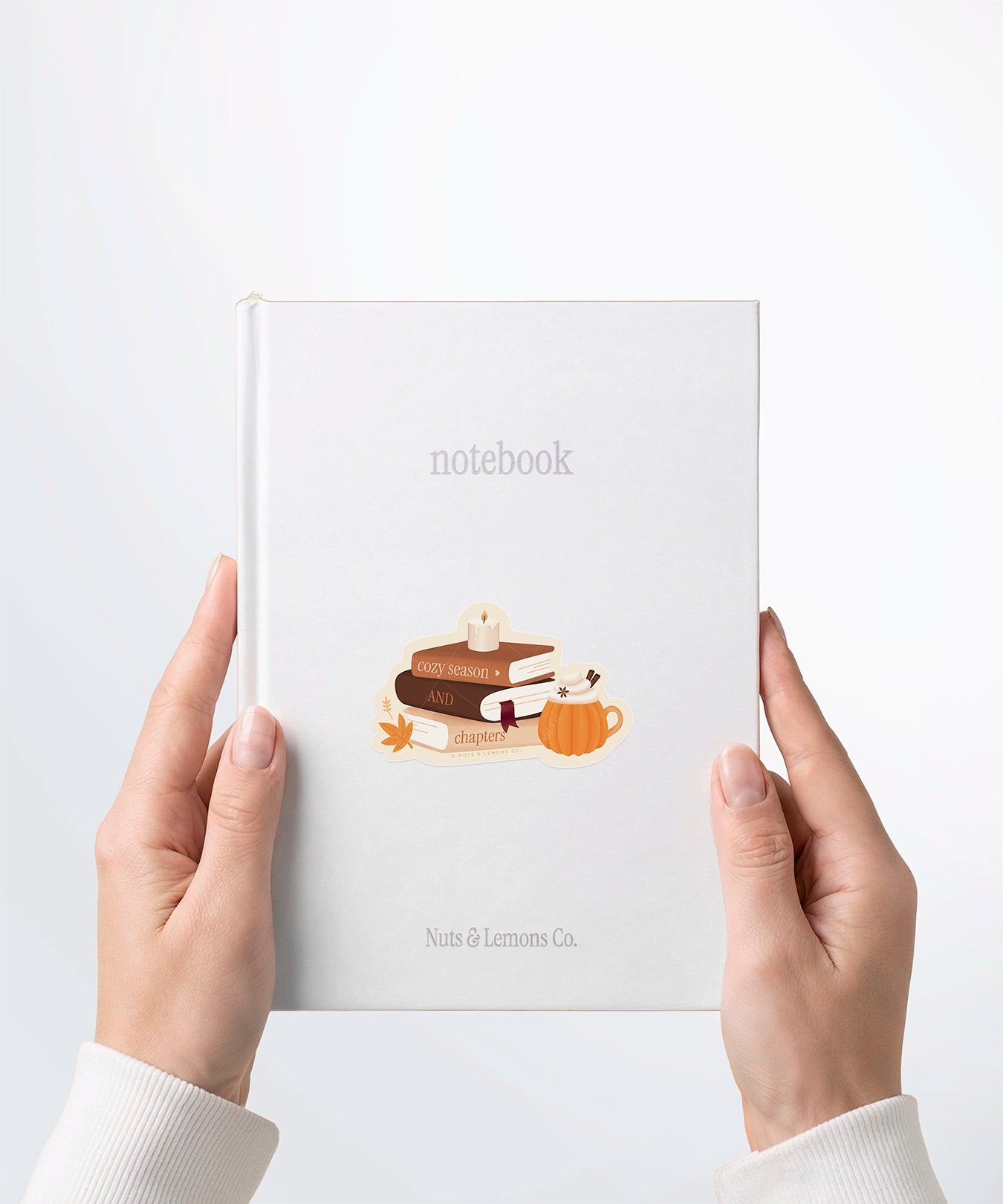 A pair of hands holding a white notebook with a Nuts & Lemons Co. diecut sticker on it featuring a stack of autumnal books, candle and pumpkin spice latte with the phrase “Cozy Season and Chapters”