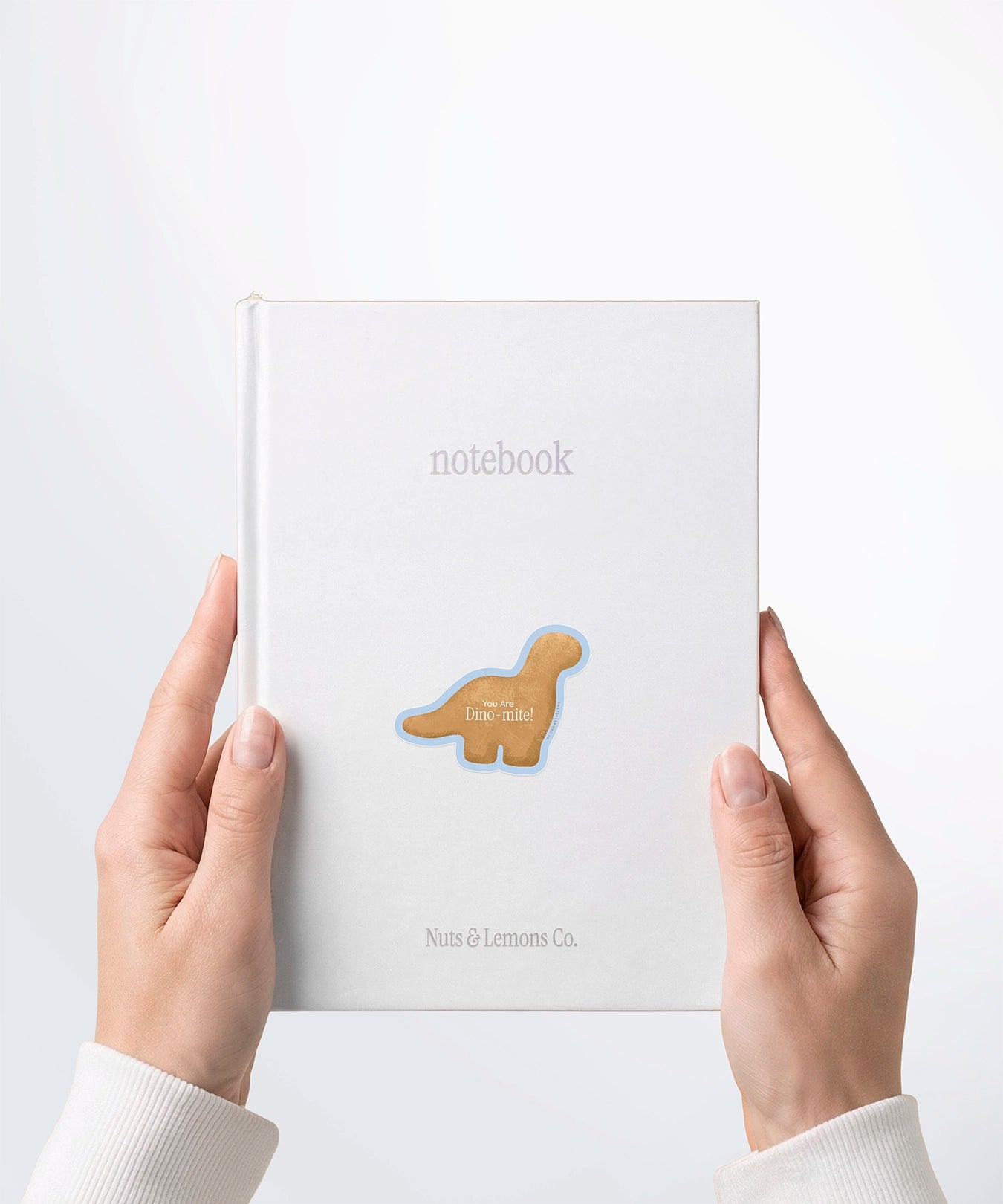 A pair of hands holding a white notebook with a Nuts & Lemons Co. diecut sticker on it featuring a Dino nug with the phrase “You Are Dino-mite”