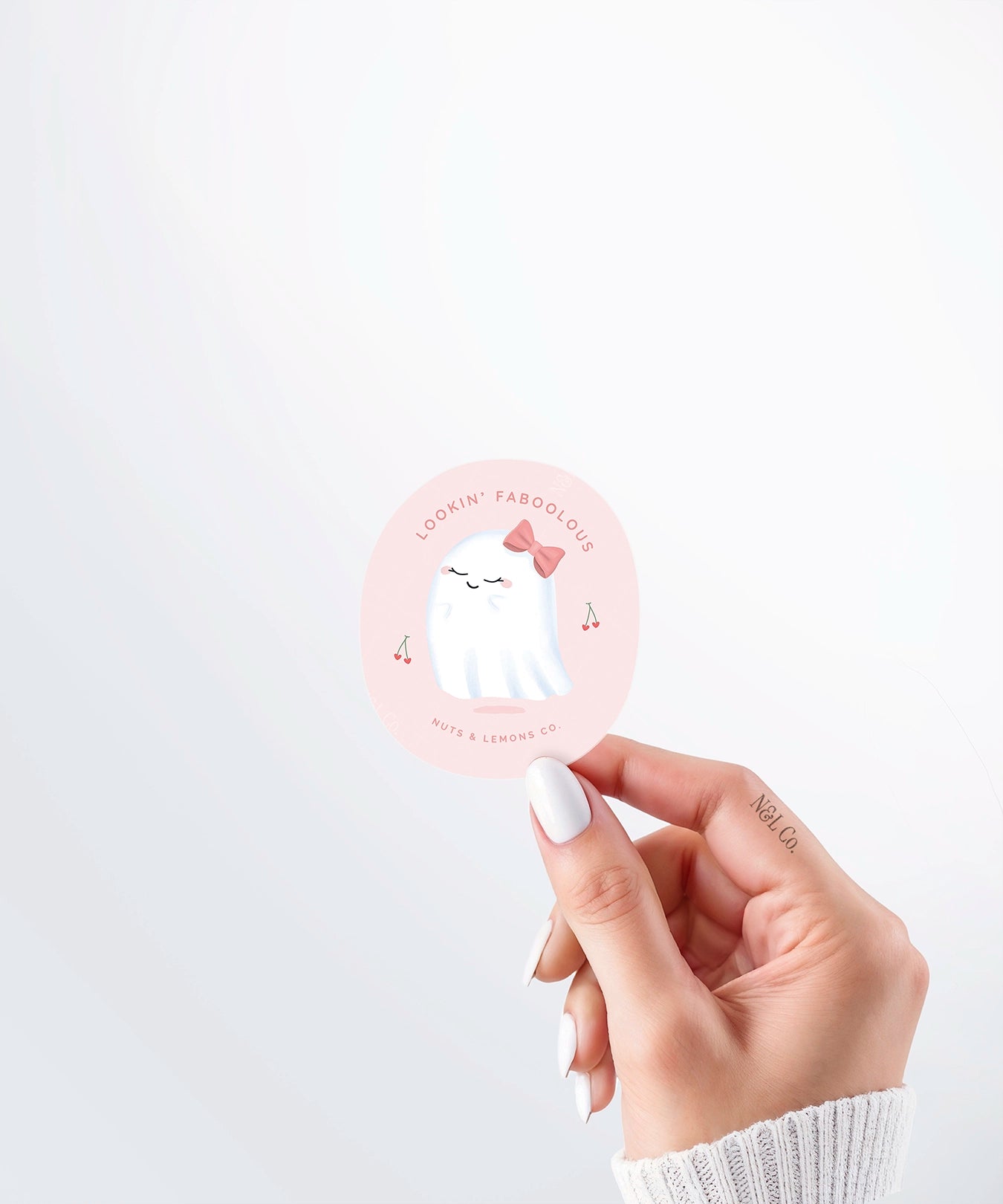 Hand holding a Nuts & Lemons Co. diecut sticker featuring a cute ghostie with a pink bow surrounded by heart cherries with the phrase “lookin’ faboolous”