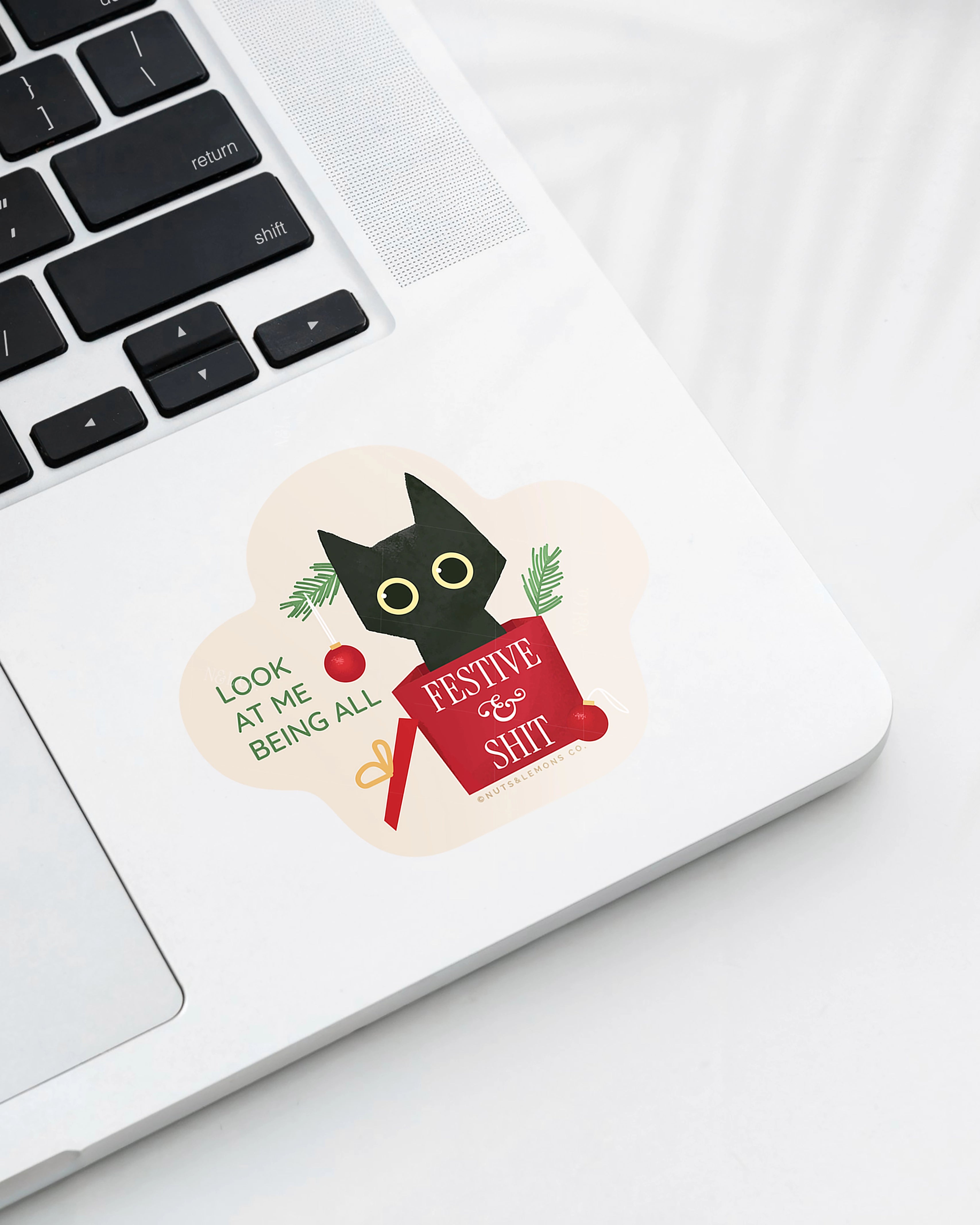 Laptop displaying a Nuts & Lemons Co. diecut sticker featuring a black cat that has rummaged thru a Christmas tree into a present with the phrase “look at me being all festive and sh*t”
