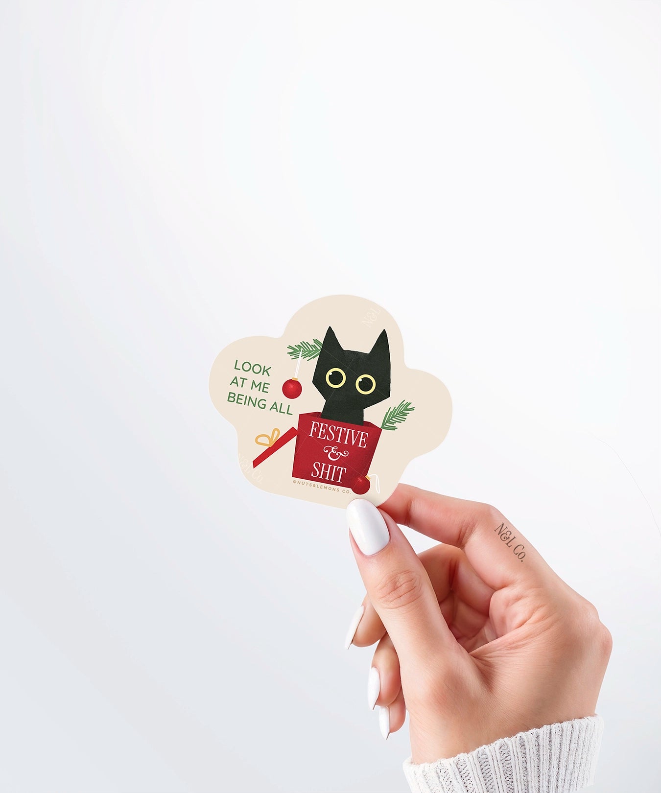 Hand holding a Nuts & Lemons Co. diecut sticker featuring a black cat that has rummaged thru a Christmas tree into a present with the phrase “look at me being all festive and sh*t”