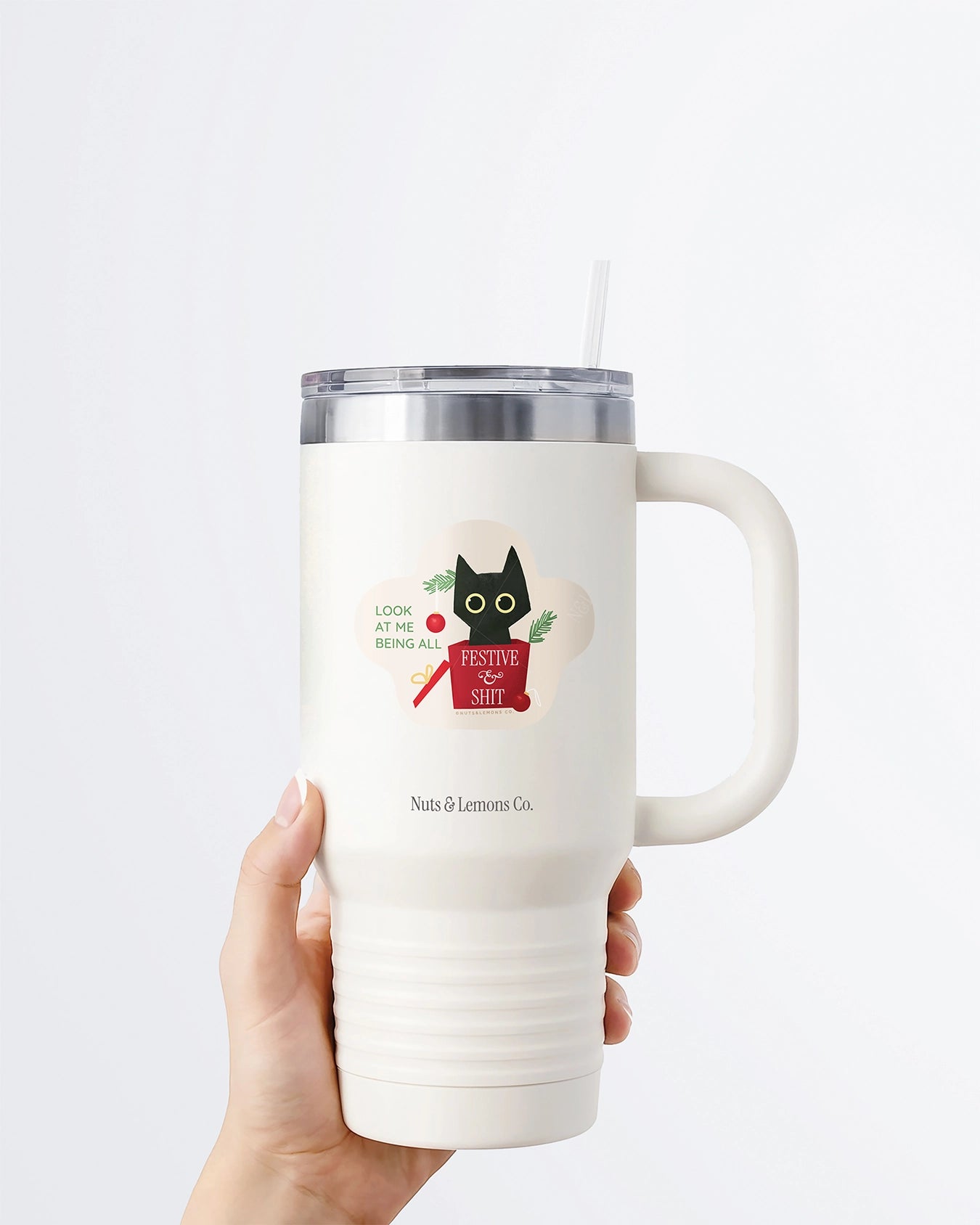 Hand holding a tumbler displaying Nuts & Lemons Co. diecut sticker featuring a black cat that has rummaged thru a Christmas tree into a present with the phrase “look at me being all festive and sh*t”