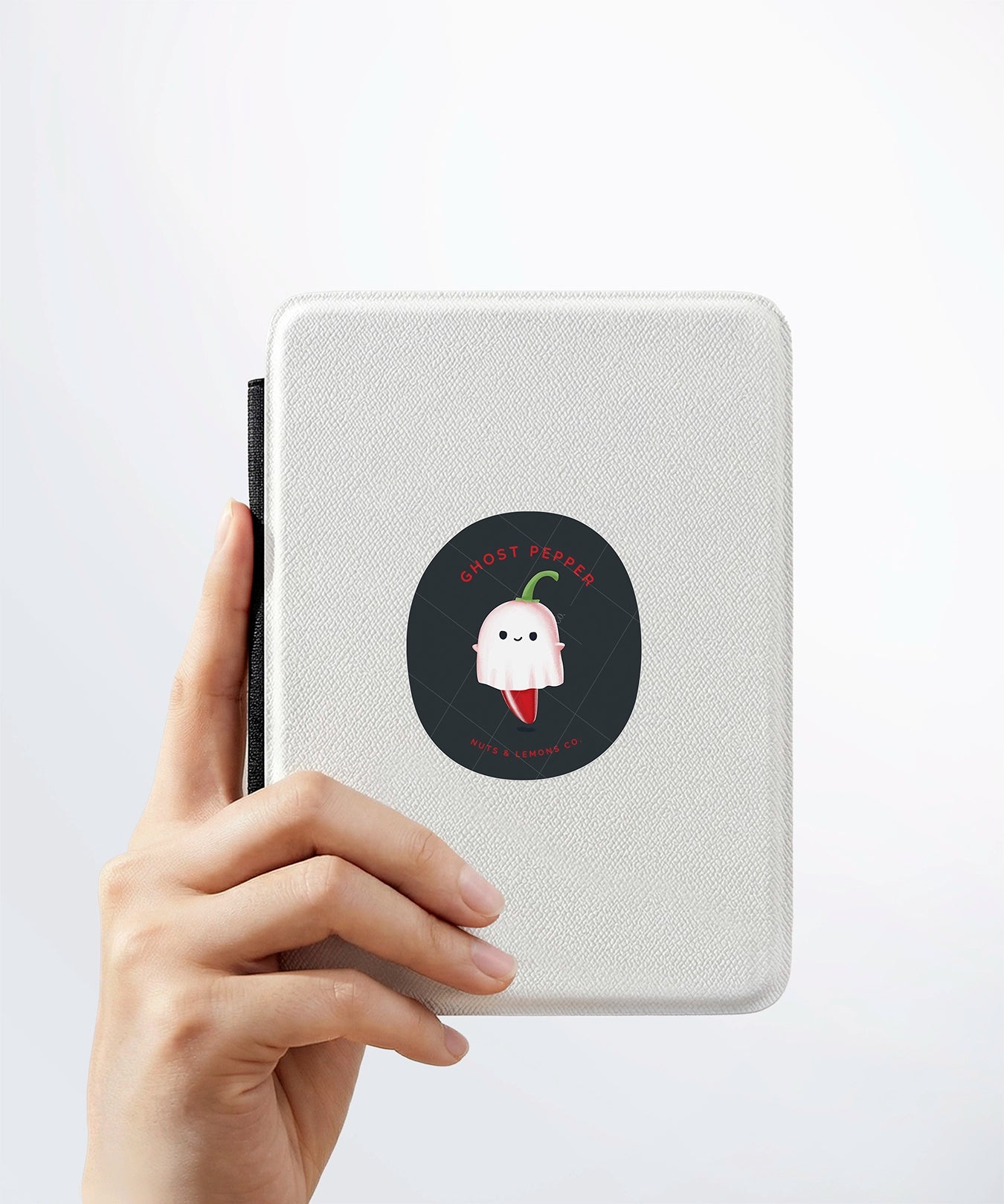 A hand holding a white kindle cover with a Nuts & Lemons Co. diecut sticker on it featuring cute ghost pepper dressed as a ghostie with the phrase “Ghost Pepper”