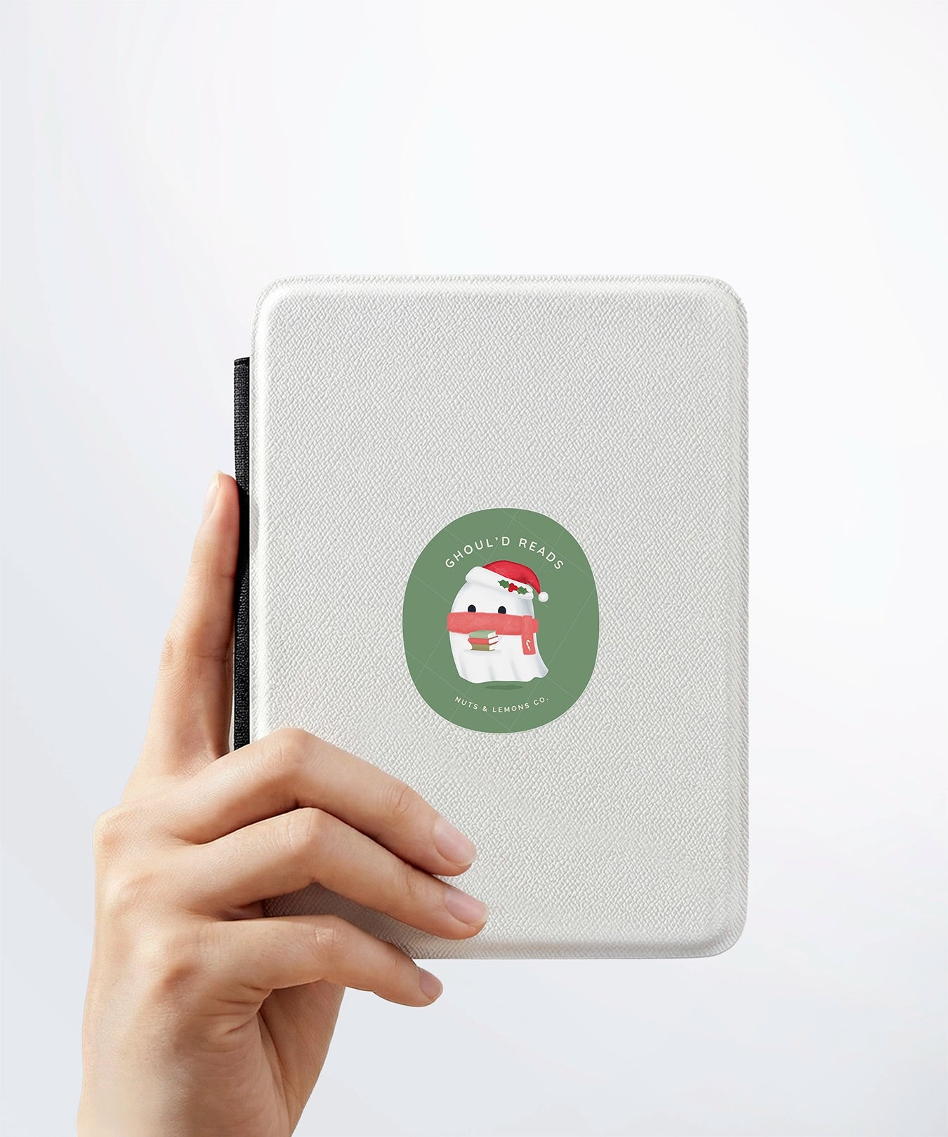 A hand holding a white kindle cover with a Nuts & Lemons Co. diecut sticker on it featuring a cute ghostie with books, Santa hat and scarf with the phrase “Ghoul’d Reads”