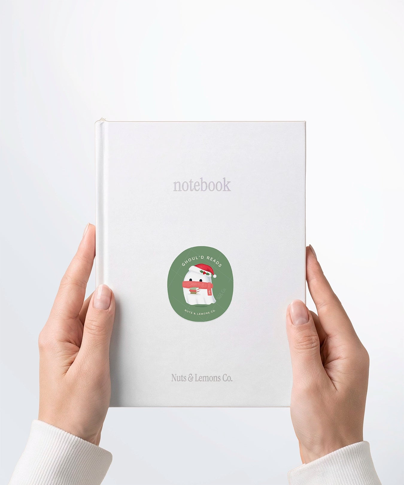 A pair of hands holding a white notebook with a Nuts & Lemons Co. diecut sticker on it featuring a cute ghostie with books, Santa hat and scarf with the phrase “Ghoul’d Reads”