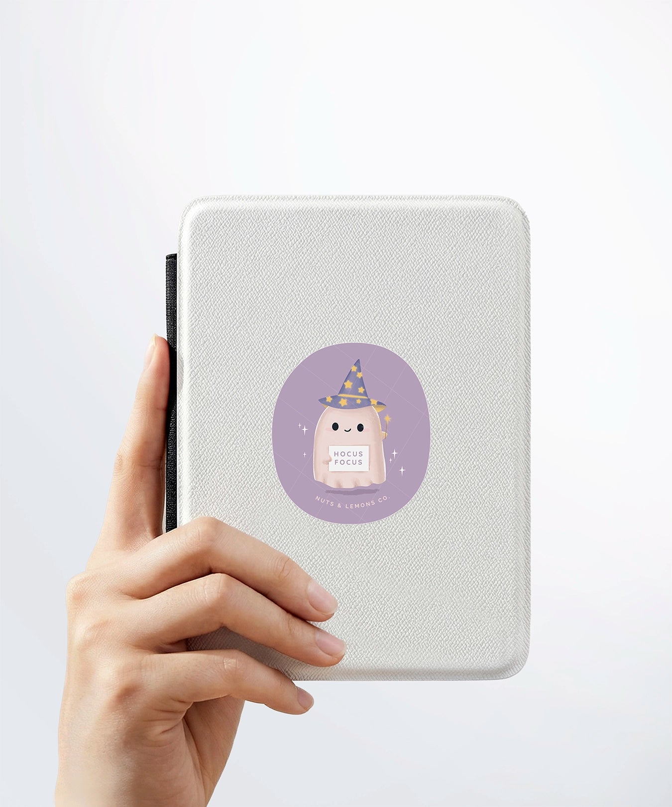 A hand holding a white kindle cover with a Nuts & Lemons Co. diecut sticker on it featuring a cute ghostie wearing a starry wizard hat, a magic wand and a sign with the phrase “Hocus Focus”