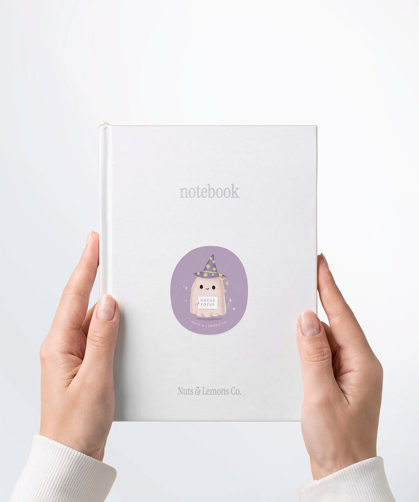 A pair of hands holding a white notebook with a Nuts & Lemons Co. diecut sticker on it featuring a cute ghostie wearing a starry wizard hat, a magic wand and a sign with the phrase “Hocus Focus”