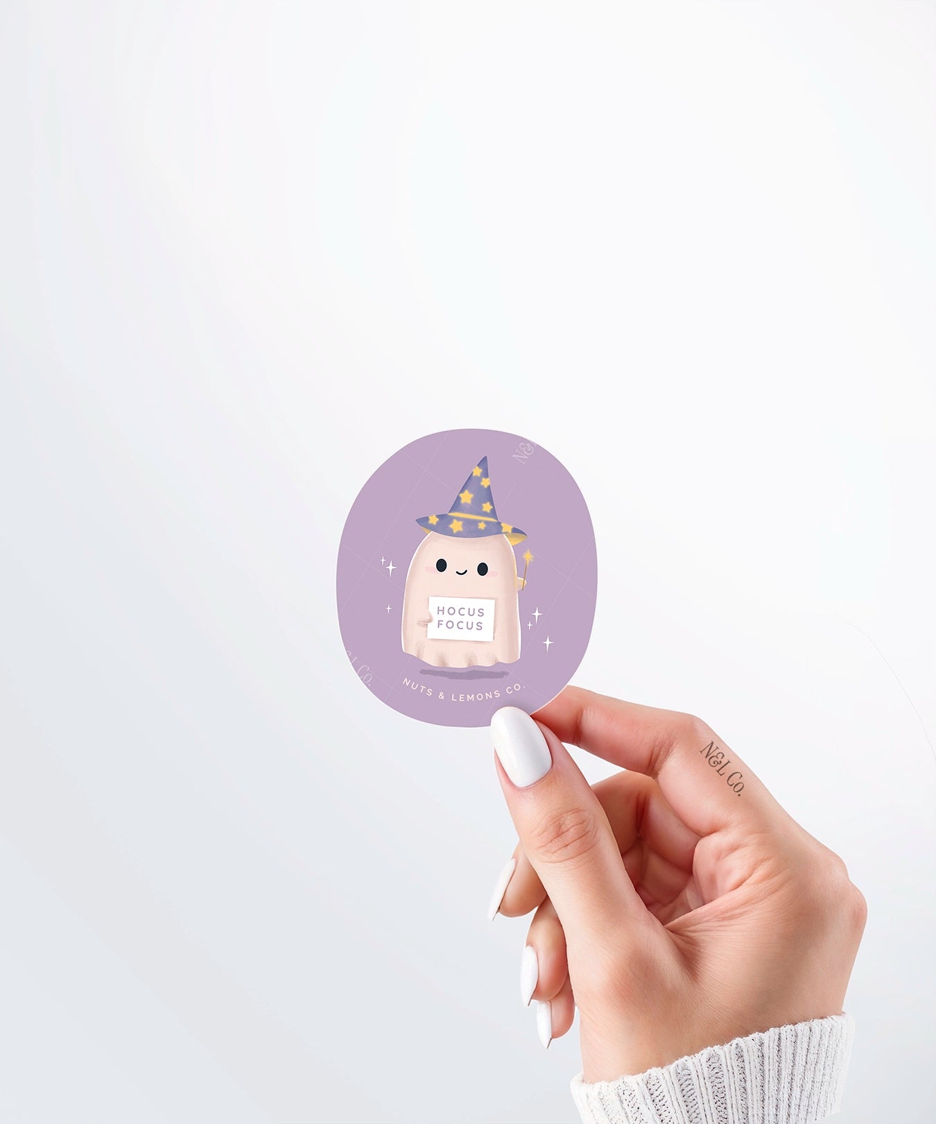 Hand holding a Nuts & Lemons Co. diecut sticker featuring a cute ghostie wearing a starry wizard hat, a magic wand  and a sign with the phrase “hocus focus”