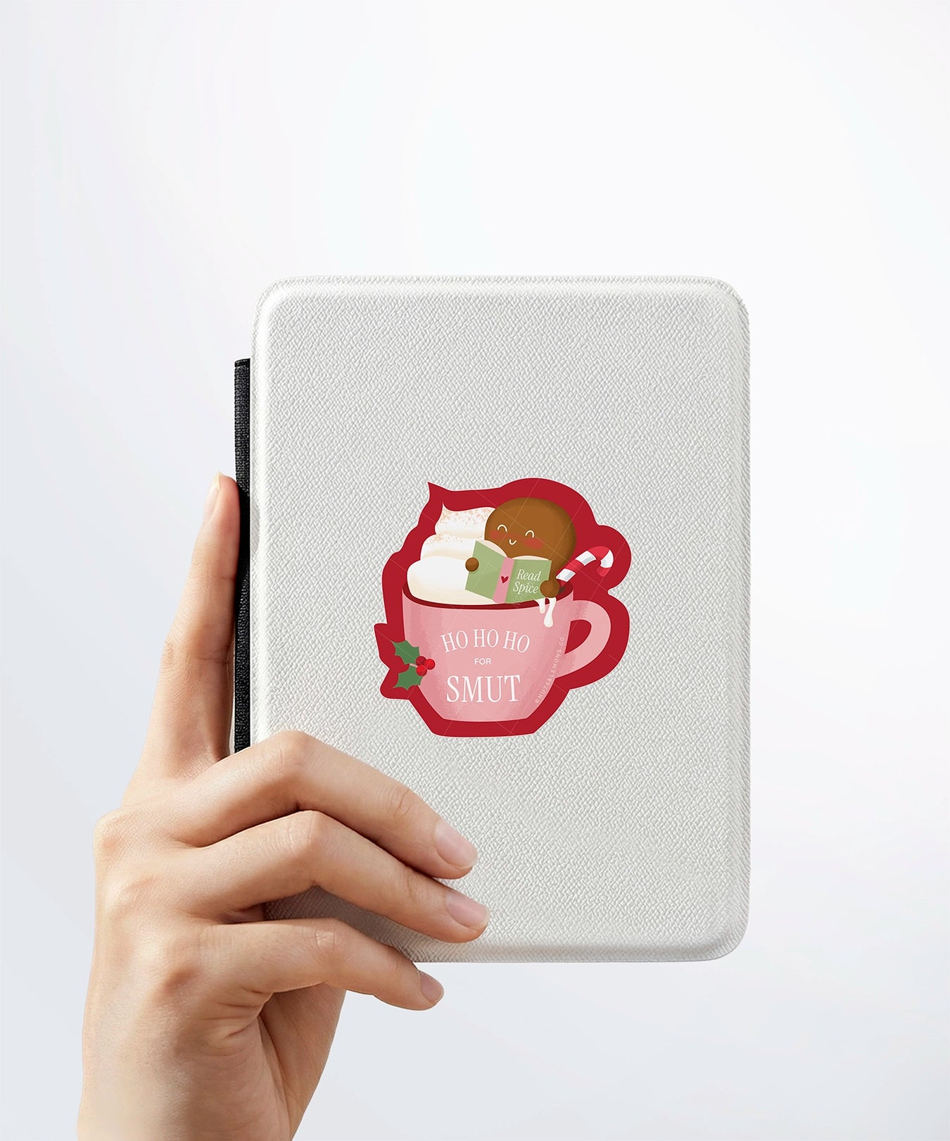 A hand holding a white kindle cover with a Nuts & Lemons Co. diecut sticker on it featuring a blushing gingerbread reading a spicy book while holding a candy cane hot tubbing in a pink mug with the phrase “Ho Ho Ho for Smut”