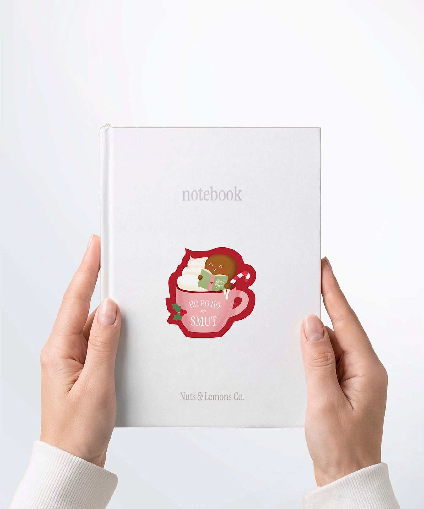 A pair of hands holding a white notebook with a Nuts & Lemons Co. diecut sticker on it featuring a blushing gingerbread reading a spicy book while holding a candy cane hot tubbing in a pink mug with the phrase “Ho Ho Ho for Smut”
