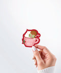 Hand holding a Nuts & Lemons Co. diecut sticker featuring a blushing gingerbread reading a spicy book while holding a candy cane hot tubbing in a pink mug with the phrase “hohoho for smut”