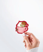 Hand holding a Nuts & Lemons Co. diecut sticker featuring a blushing gingerbread reading a spicy book while holding a candy cane hot tubbing in a pink mug with the phrase “hohoho for smut”