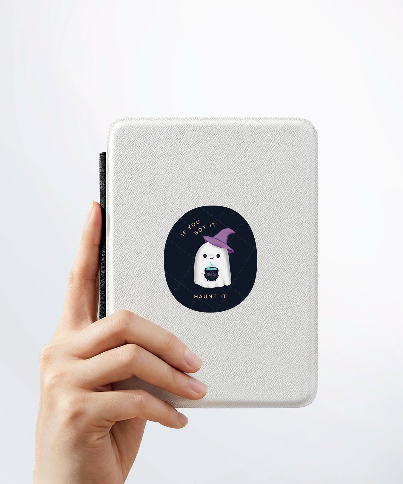 A hand holding a white kindle cover with a Nuts & Lemons Co. diecut sticker on it featuring a cute ghostie in a witch hat with a bubbling cauldron with the phrase “If You Got It, Haunt It”