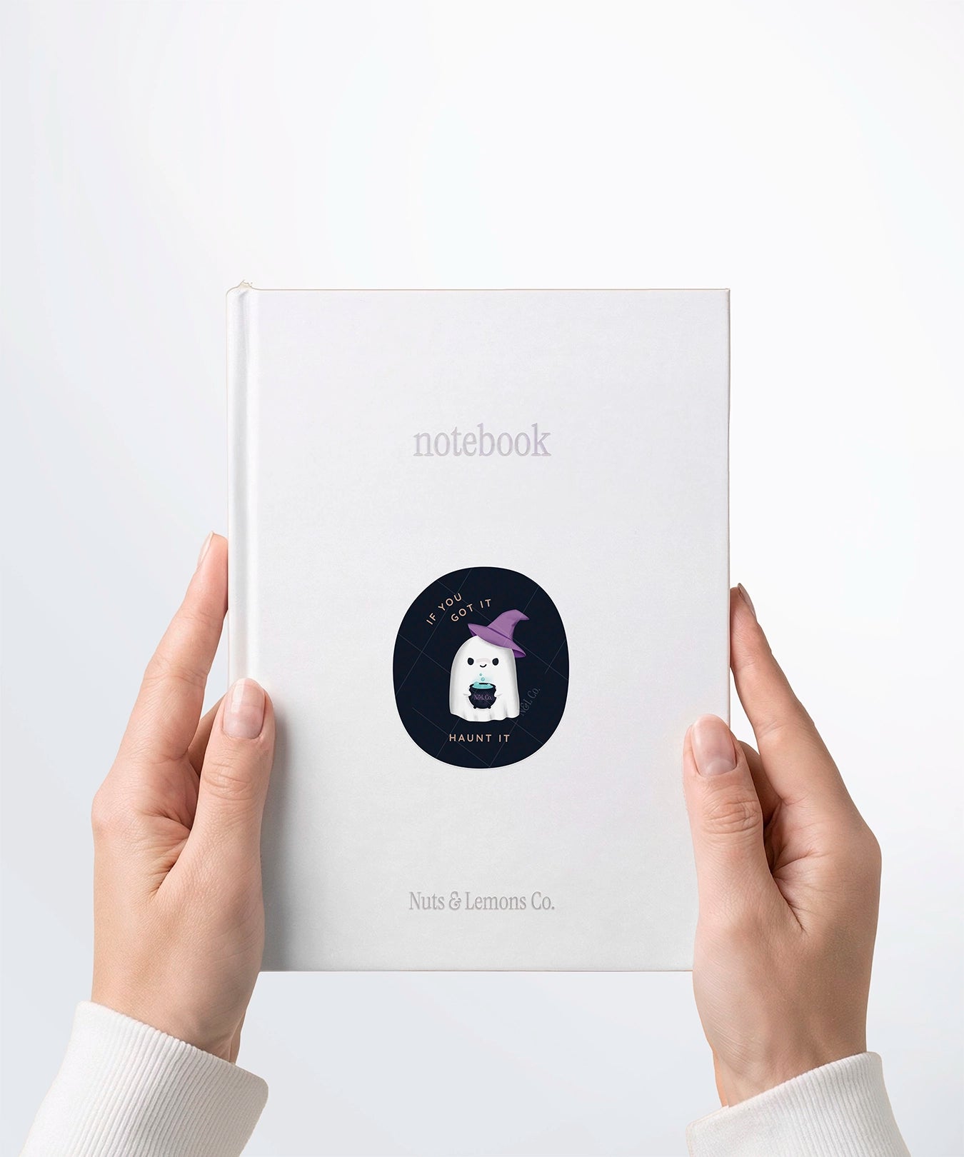 A pair of hands holding a white notebook with a Nuts & Lemons Co. diecut sticker on it featuring a cute ghostie in a witch hat with a bubbling cauldron with the phrase “If You Got It, Haunt It”