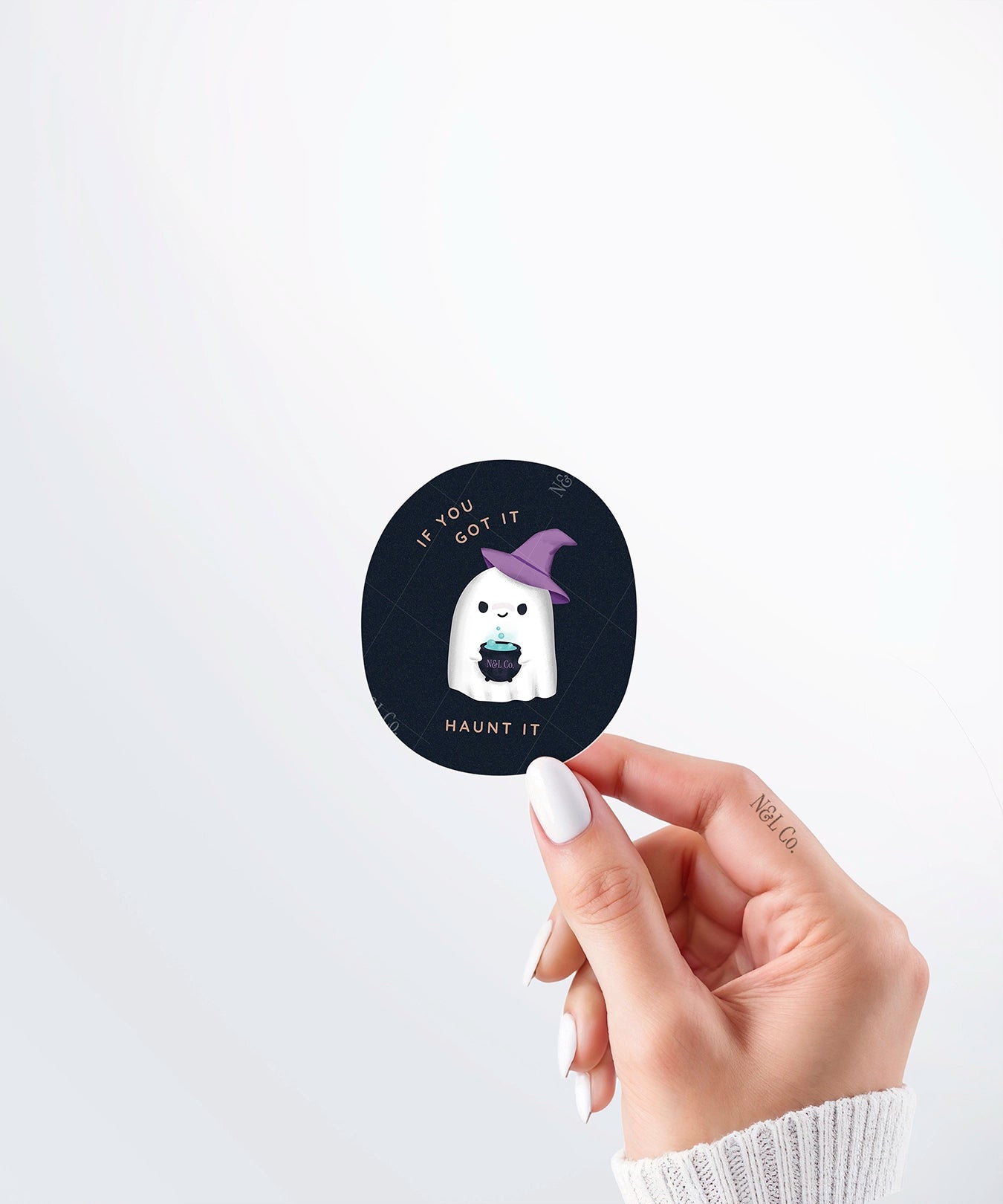 Hand holding a Nuts & Lemons Co. diecut sticker featuring a cute ghostie in a witch hat with a bubbling cauldron with the phrase “if you got it, haunt it”