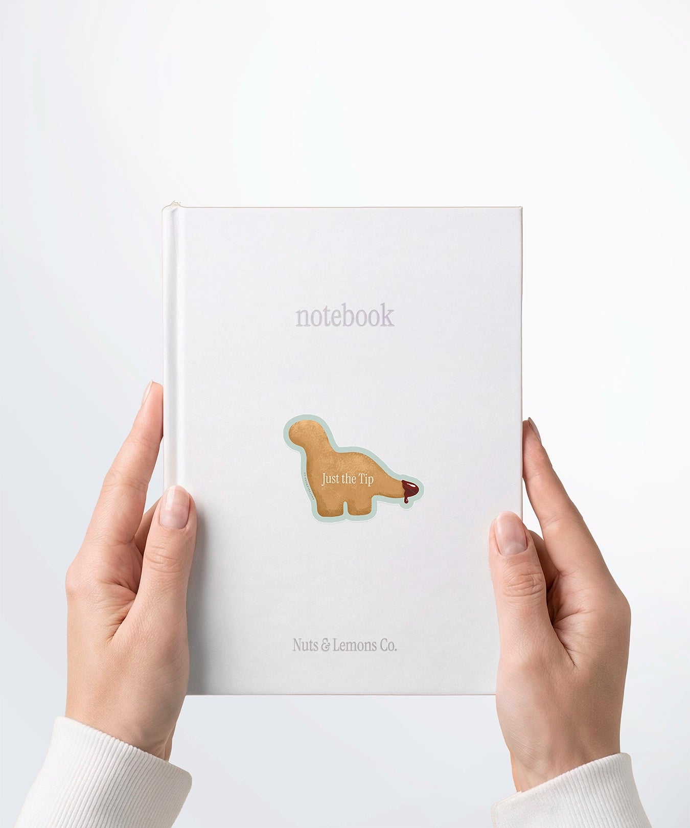 A pair of hands holding a white notebook with a Nuts & Lemons Co. diecut sticker on it featuring a Dino nug with tail dipped in sauce with the phrase “Just The Tip”