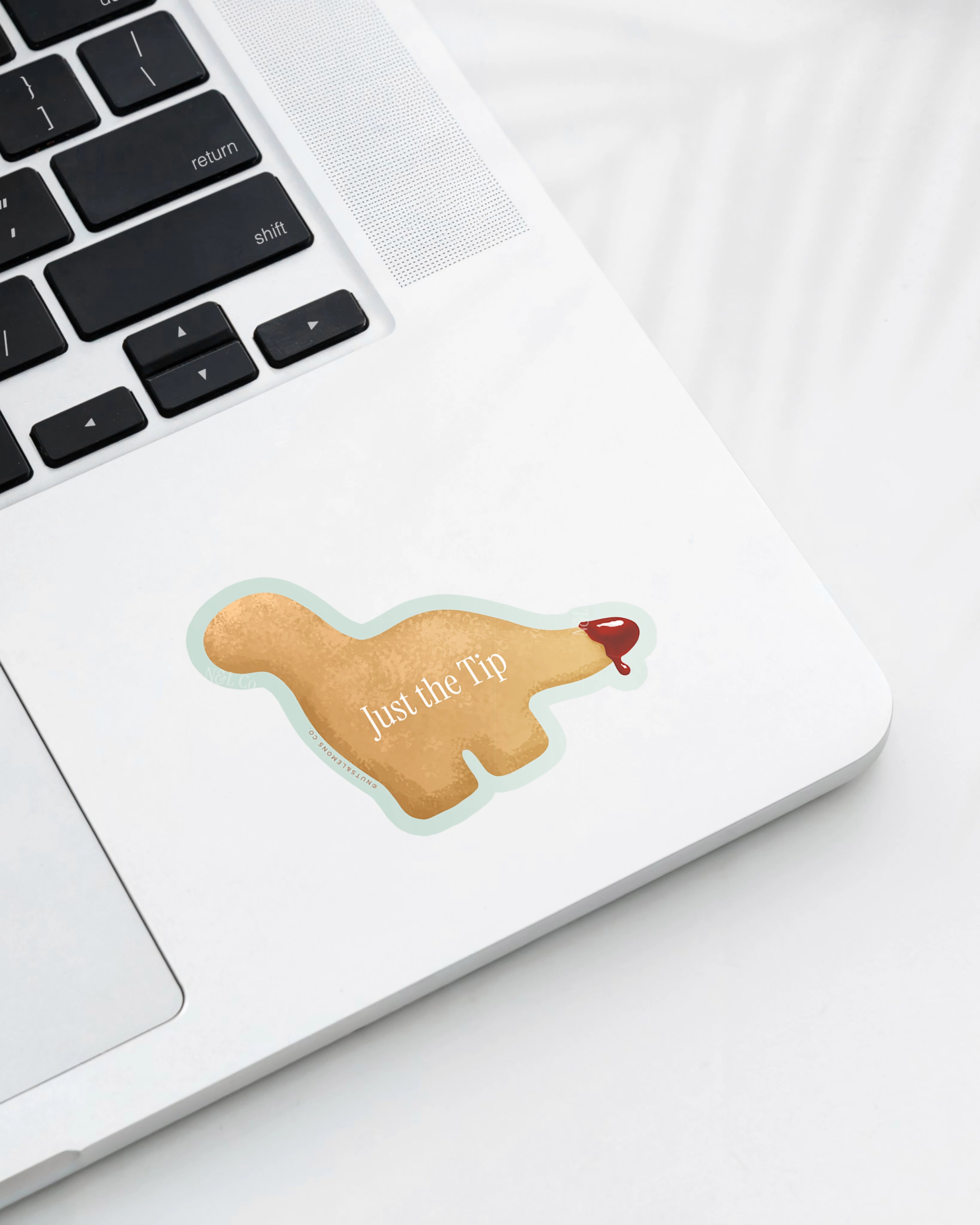 Laptop displaying a Nuts & Lemons Co. diecut sticker featuring a Dino nug with tail dipped in sauce with the phrase “just the tip”