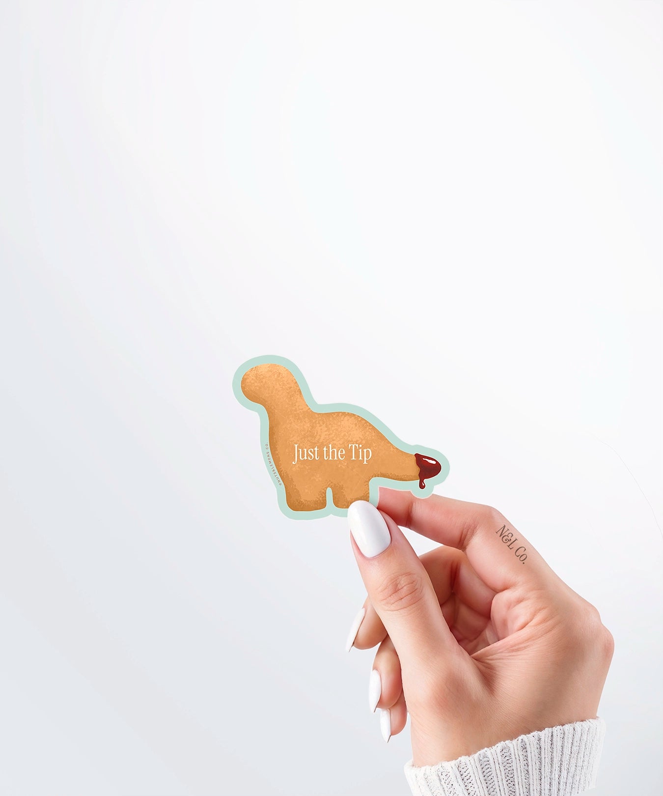Hand holding a Nuts & Lemons Co. diecut sticker featuring a Dino nug with tail dipped in sauce with the phrase “just the tip”
