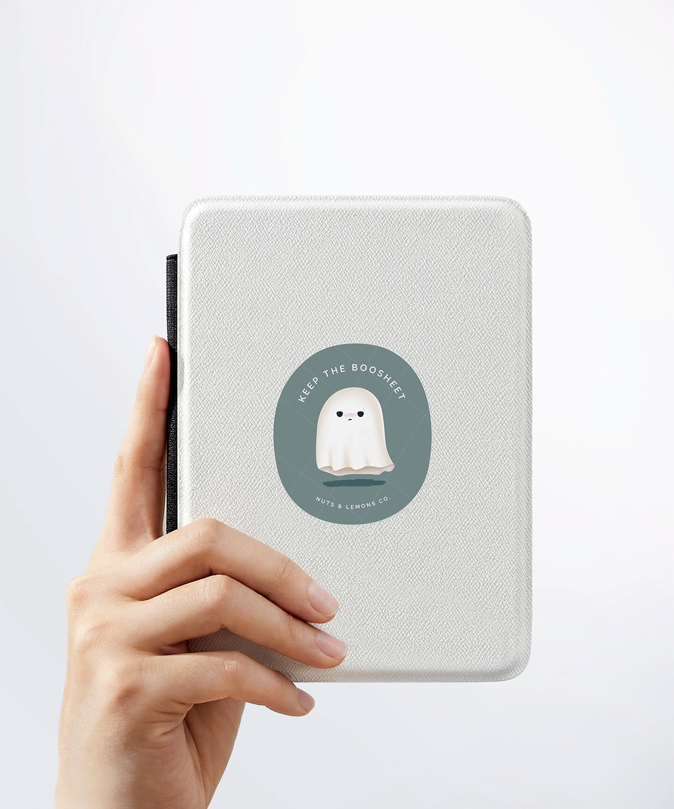 A hand holding a white kindle cover with a Nuts & Lemons Co. diecut sticker on it featuring a cute annoyed ghostie with the phrase “Keep the Boo-Sheet”