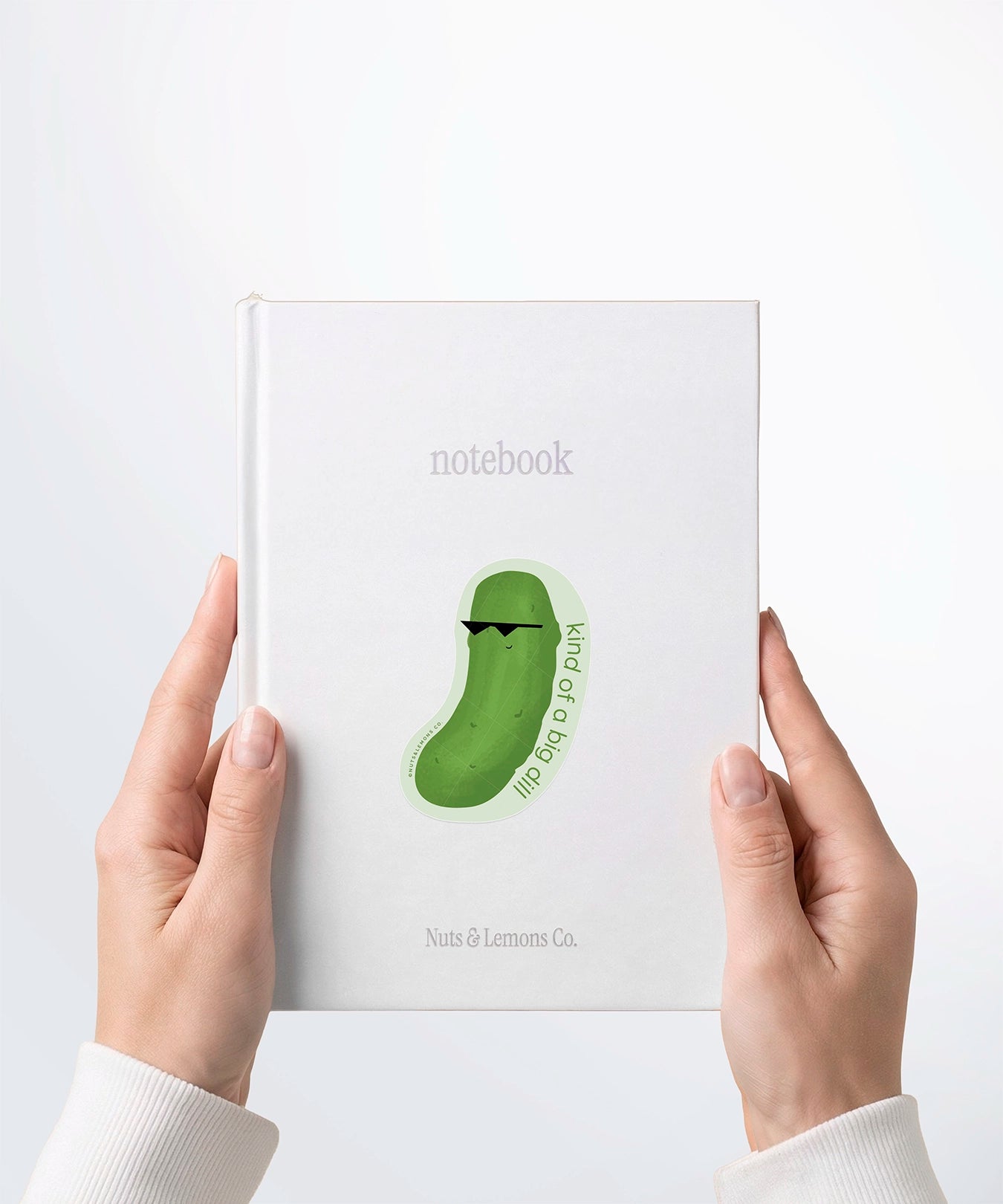 A pair of hands holding a white notebook with a Nuts & Lemons Co. diecut sticker on it featuring a dill pickle with shades with the phrase “Kind of a Big Dill”