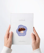 A pair of hands holding a white notebook with a Nuts & Lemons Co. diecut sticker on it featuring a calm shiitake mushroom with the phrase “Let That Shittake Go”