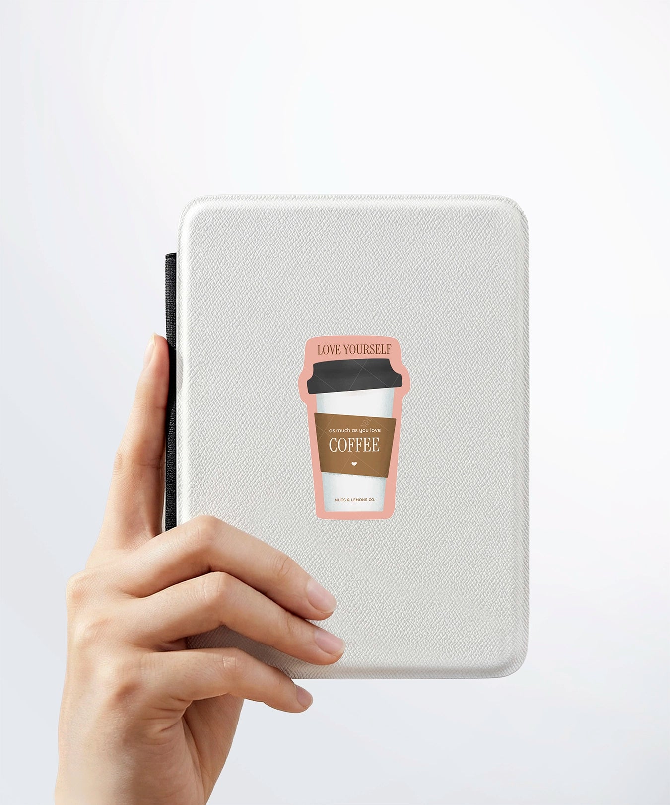 A hand holding a white kindle cover with a Nuts & Lemons Co. diecut sticker on it featuring a togo coffee with the phrase with the phrase “Love Yourself as Much as You Love Coffee”