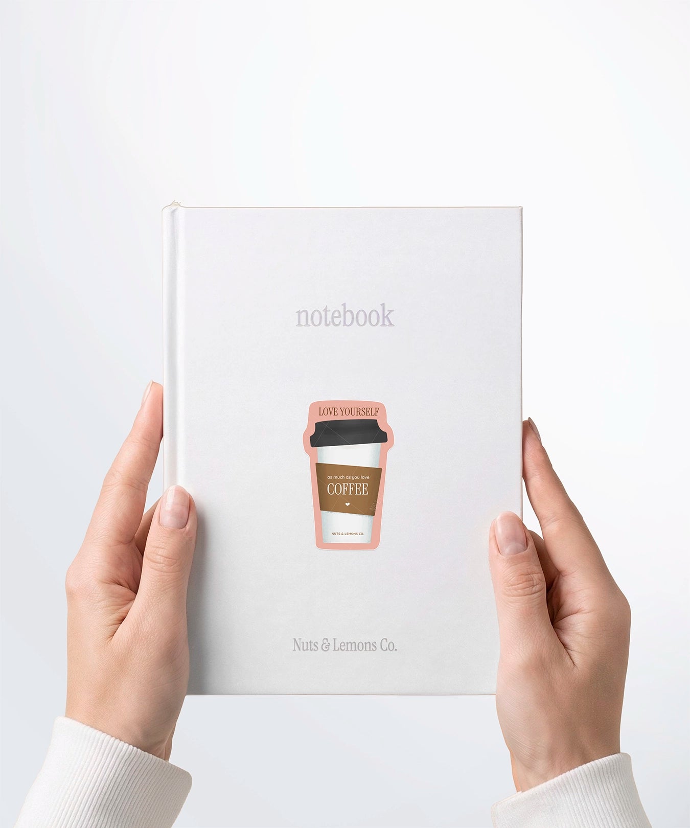 A pair of hands holding a white notebook with a Nuts & Lemons Co. diecut sticker on it featuring a togo coffee with the phrase with the phrase “Love Yourself as Much as You Love Coffee”